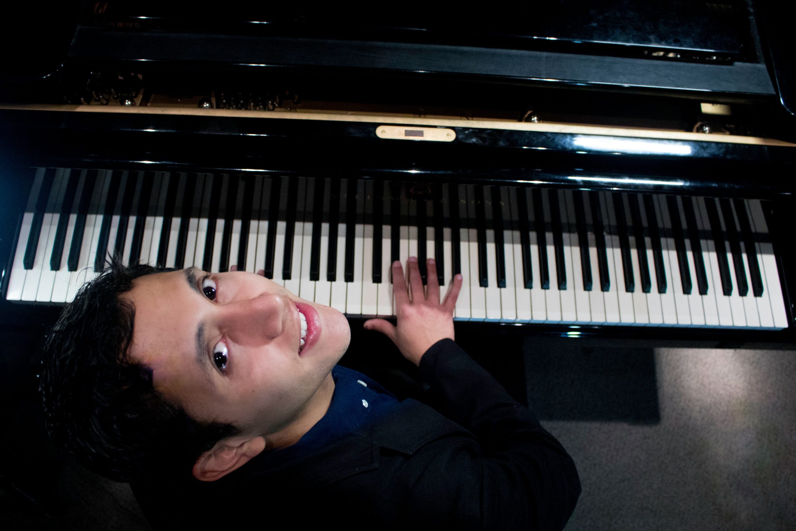 Child playing piano, looking up at camera, smiling, black hair, dark clothing, piano keys visible from above.