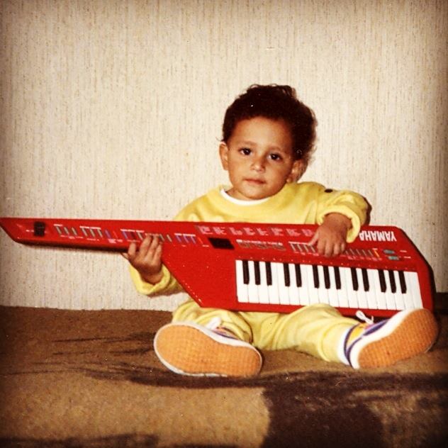 Child sitting on a carpet holding a red electronic keyboard, looking at the camera, with a plain wall background.