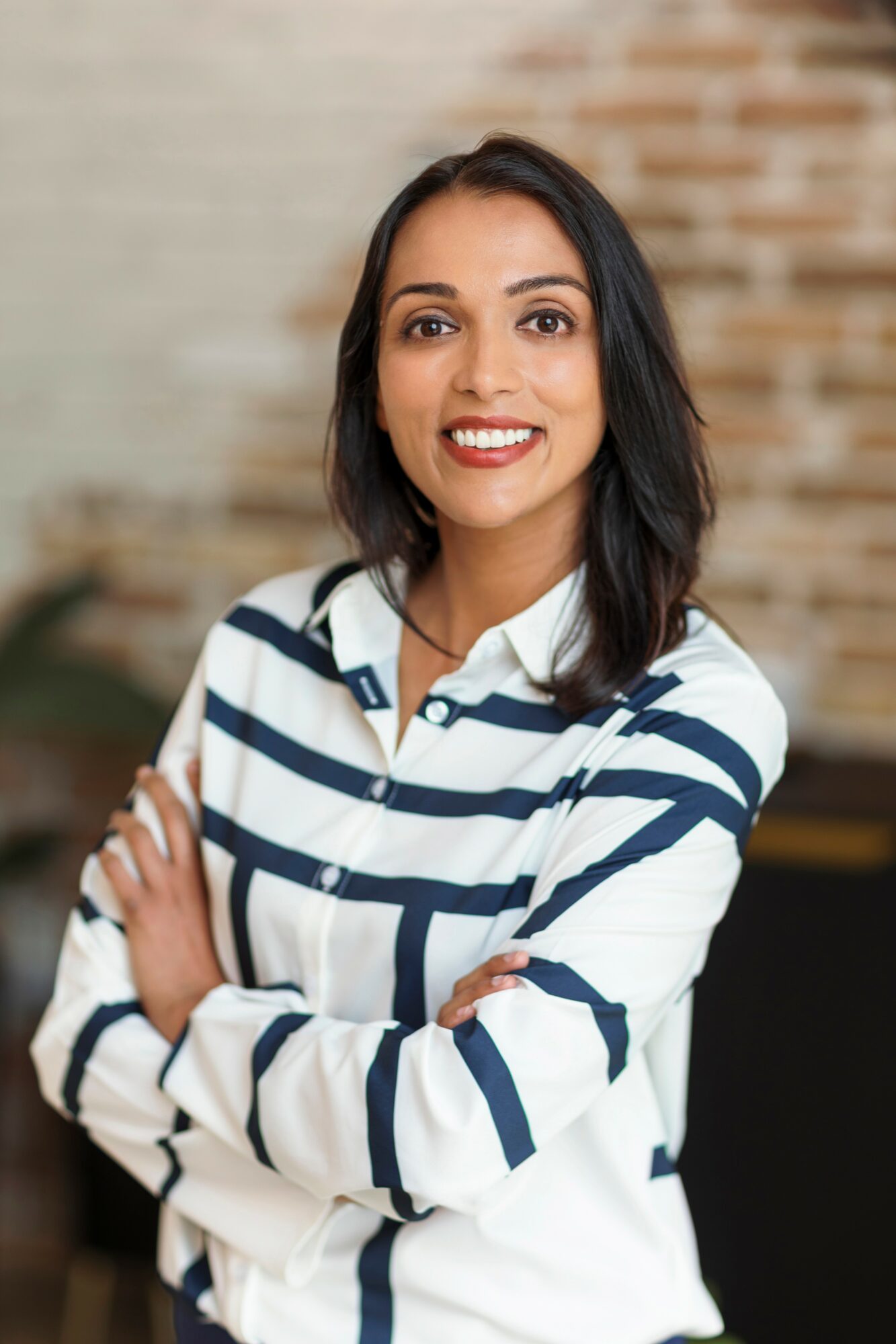 Woman with dark hair smiling, wearing a striped shirt, arms crossed, in a room with brick wall background.