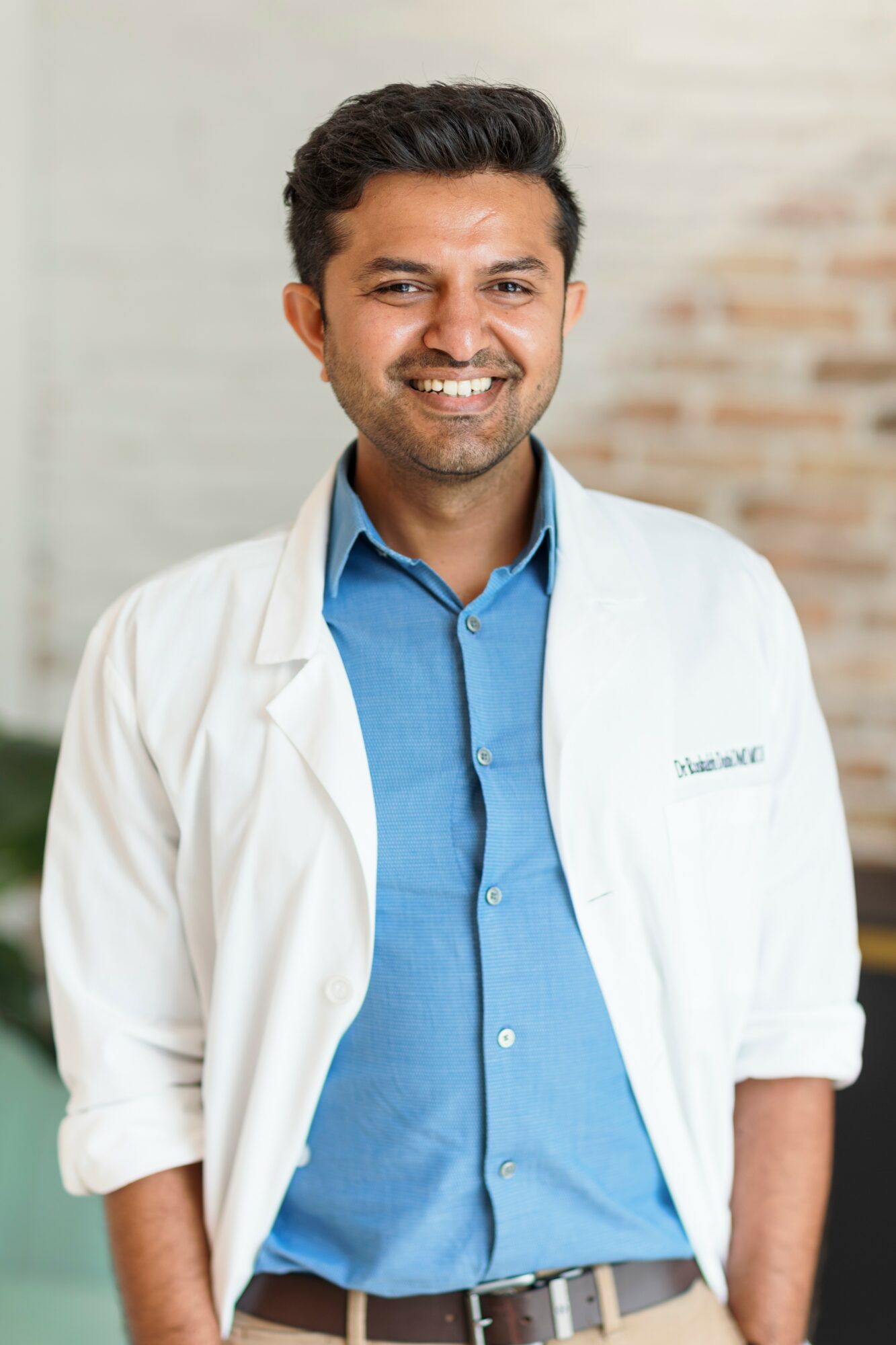 Smiling man in a white coat and blue shirt standing indoors with a brick wall background.