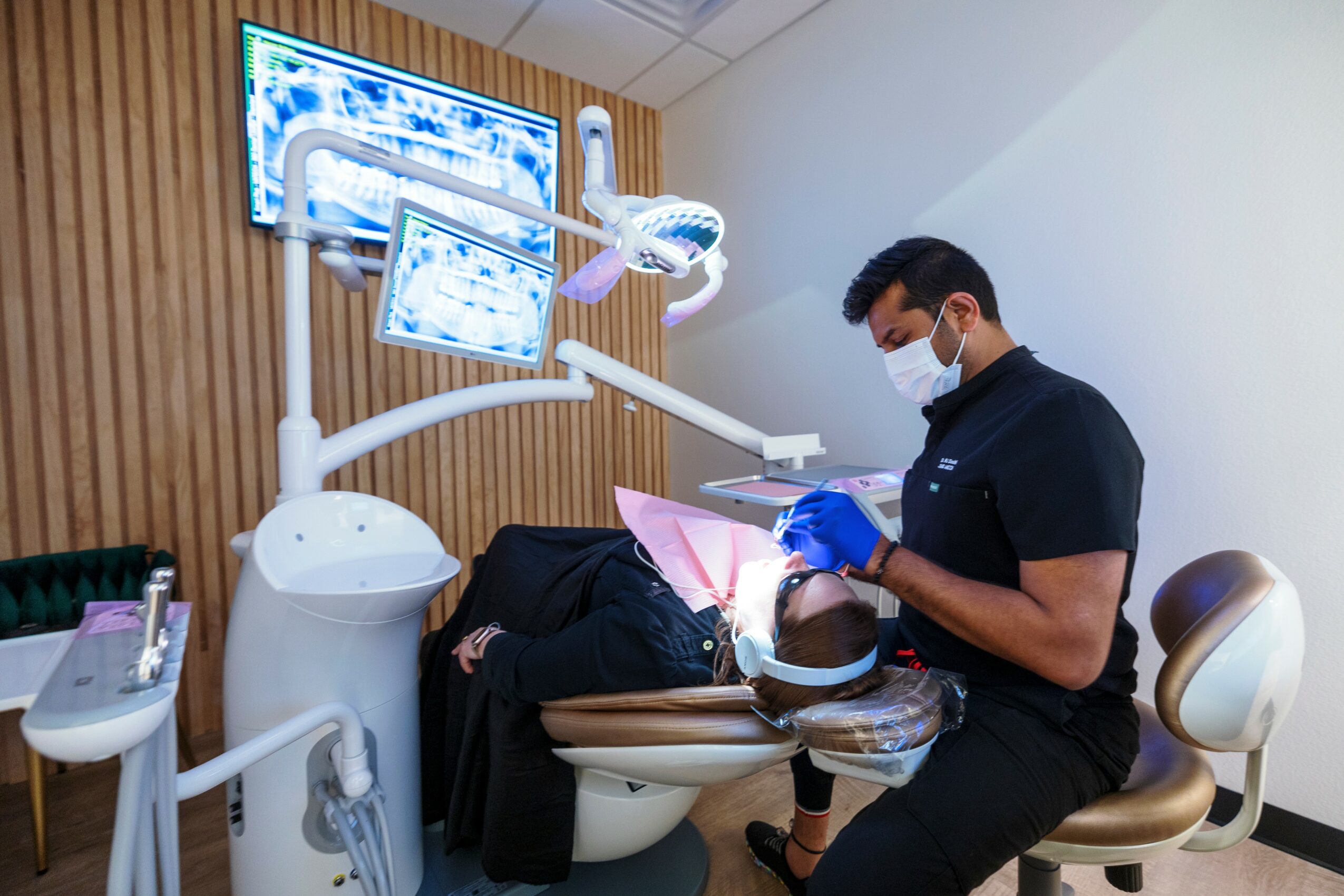 Dentist in mask and gloves working on a patient lying in dental chair, with dental equipment and monitors above. Word count: 20