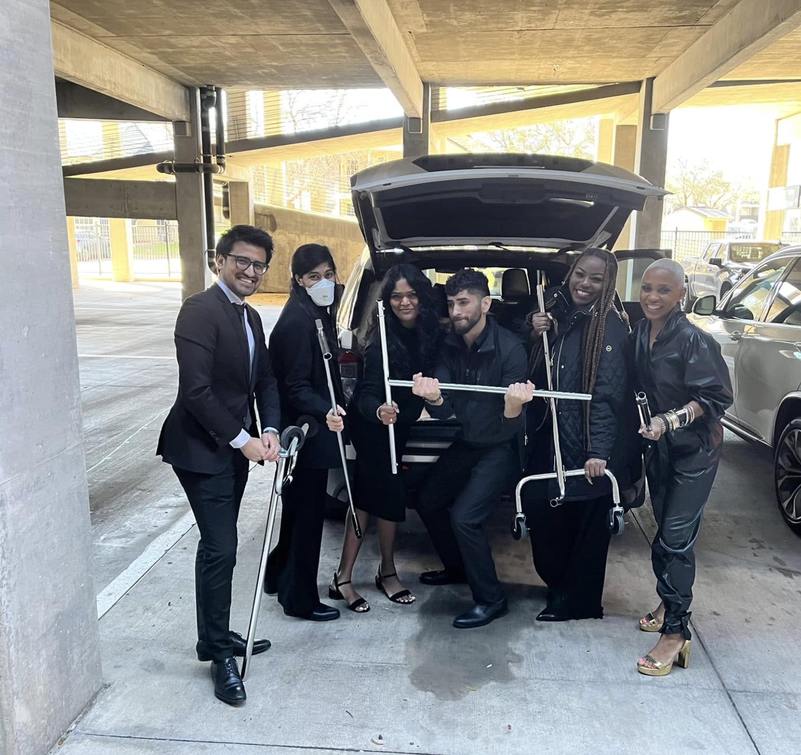 Group of six people in formal attire with canes, standing near an open car trunk in a parking garage.