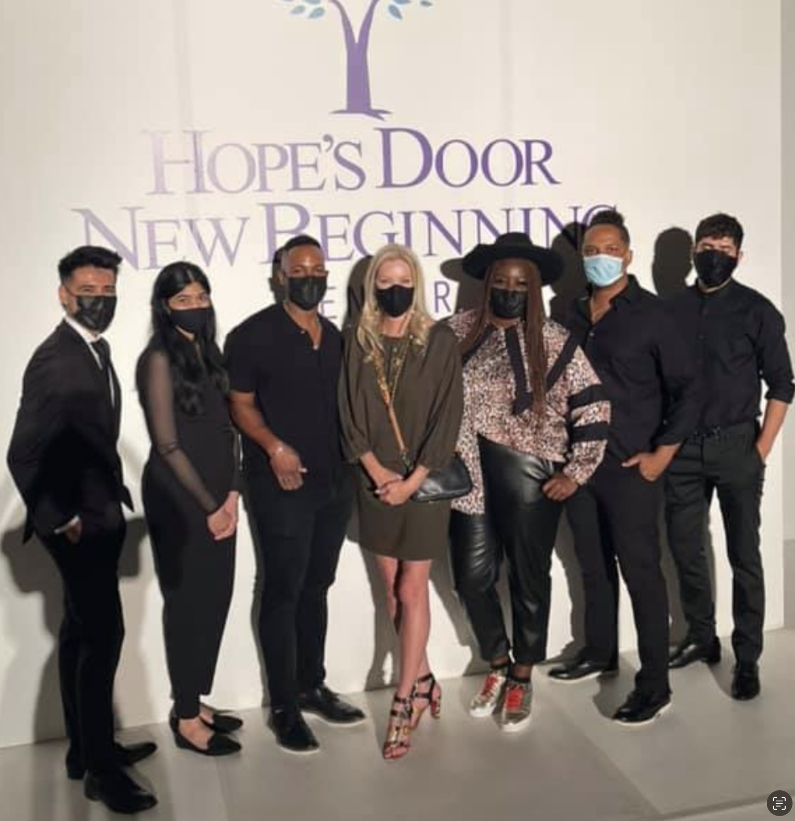 Group of eight people standing together, wearing masks, in front of a wall with a tree and text, posing for a photo.