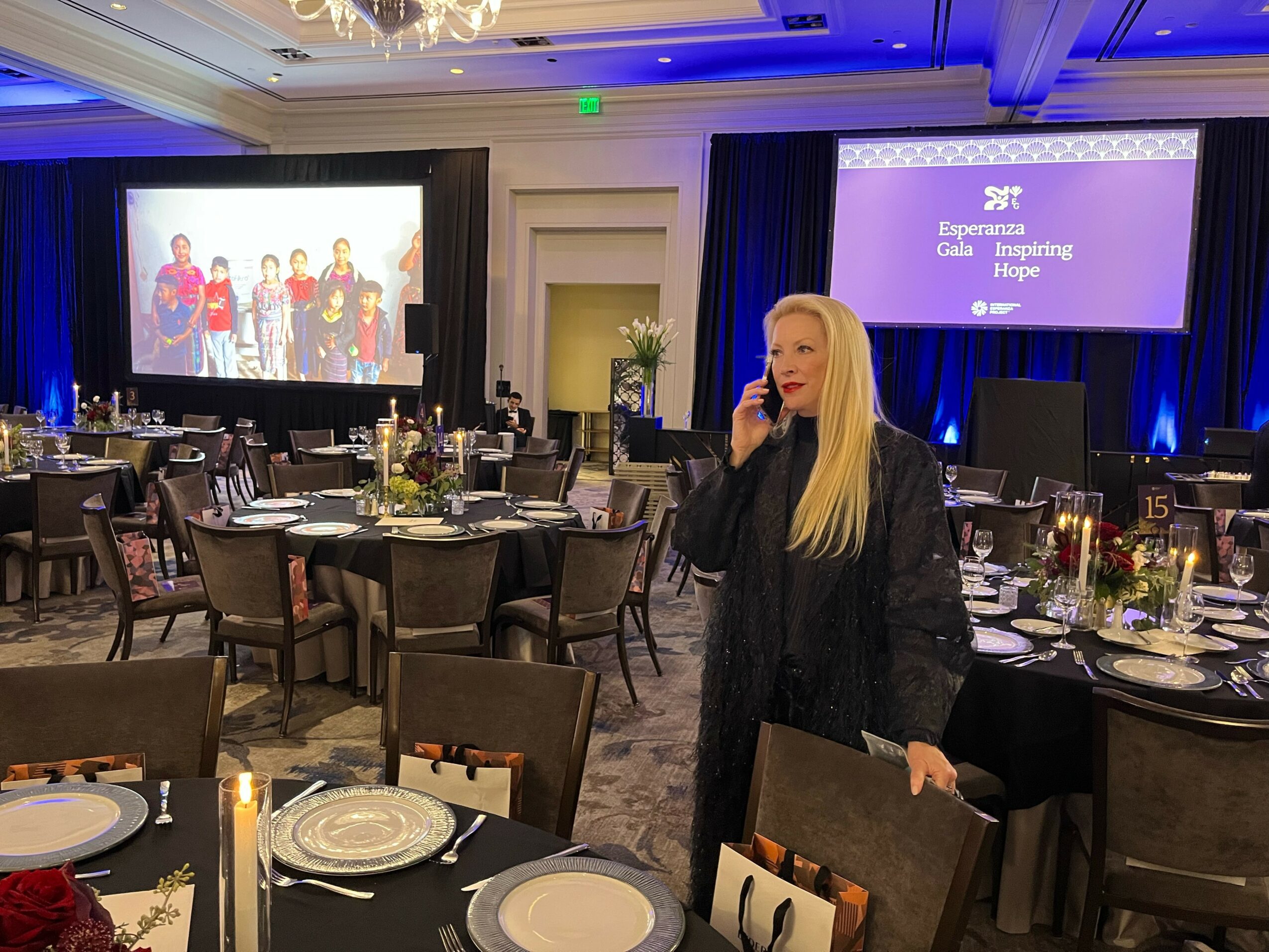 A woman with long blonde hair stands in a banquet hall, talking on her phone, with tables set for an event in the background.