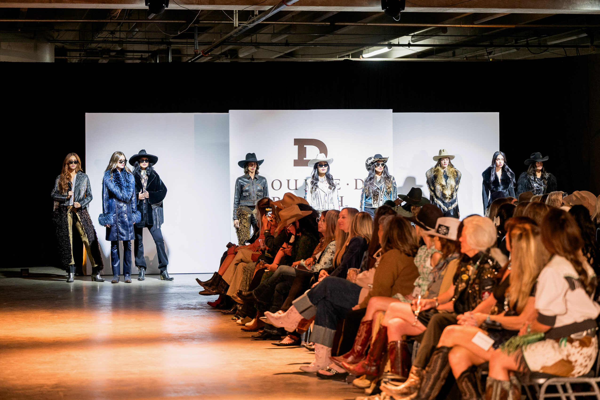 Models walk on runway with audience seated in front, backdrop displays a logo and text, in a fashion show setting.