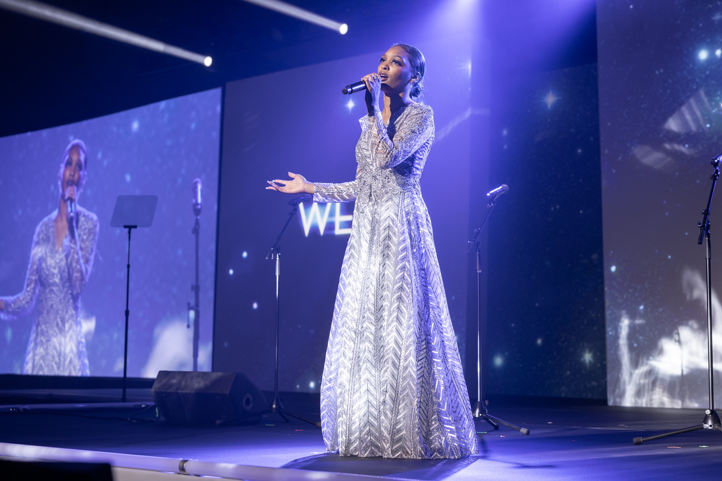 A woman in a long, patterned dress sings into a microphone on stage with large screens behind her displaying her reflection.