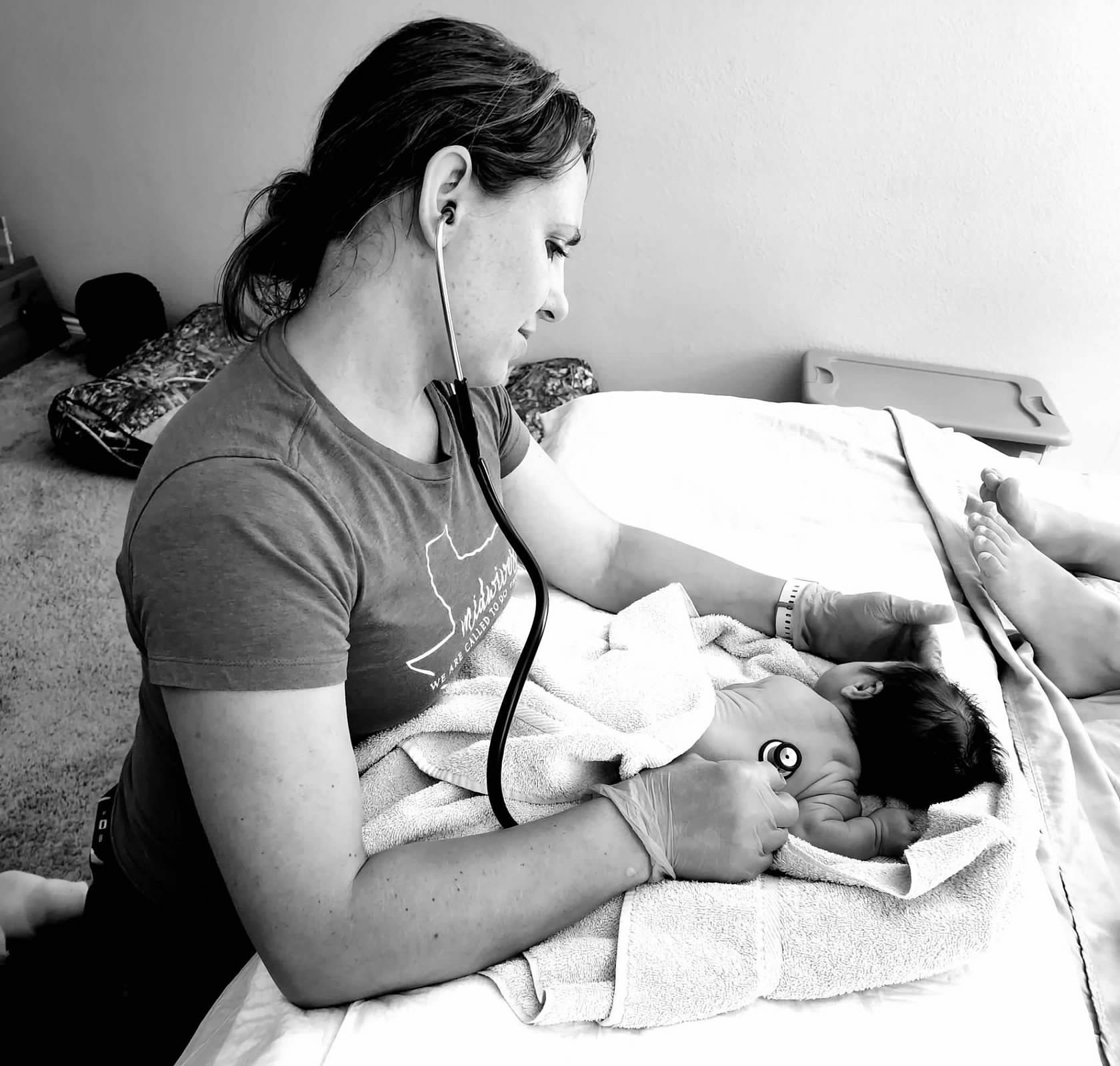 A woman with a stethoscope examines a newborn baby lying on a hospital bed, holding the baby gently.