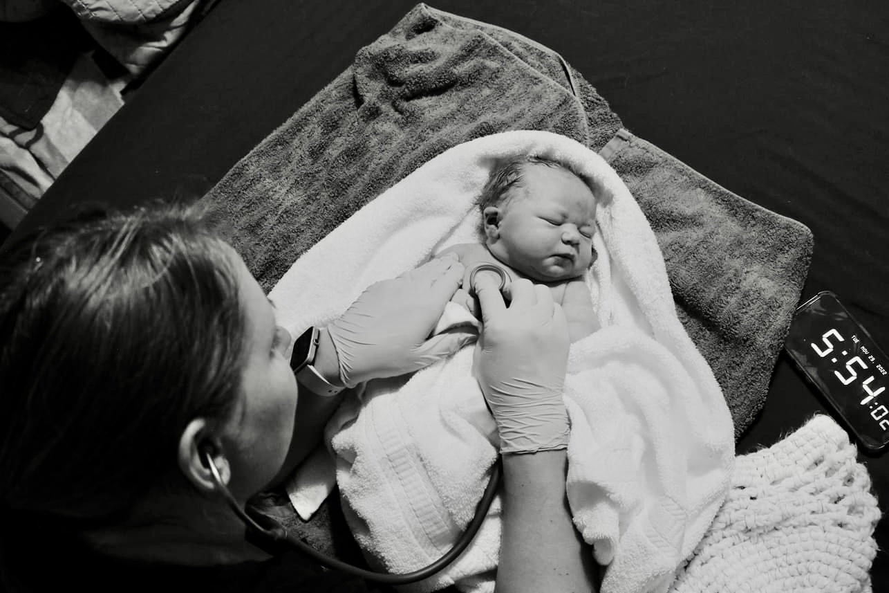 A woman holds a newborn baby wrapped in a blanket on a bed with a towel underneath, in black and white.