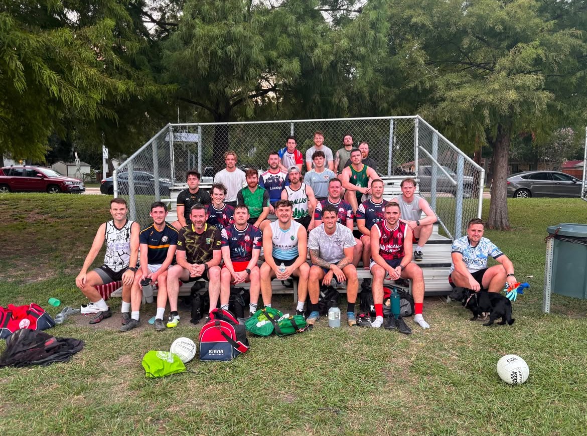 Group of people sitting on bleachers outdoors with trees and cars in background, sports bags and a soccer ball in front.