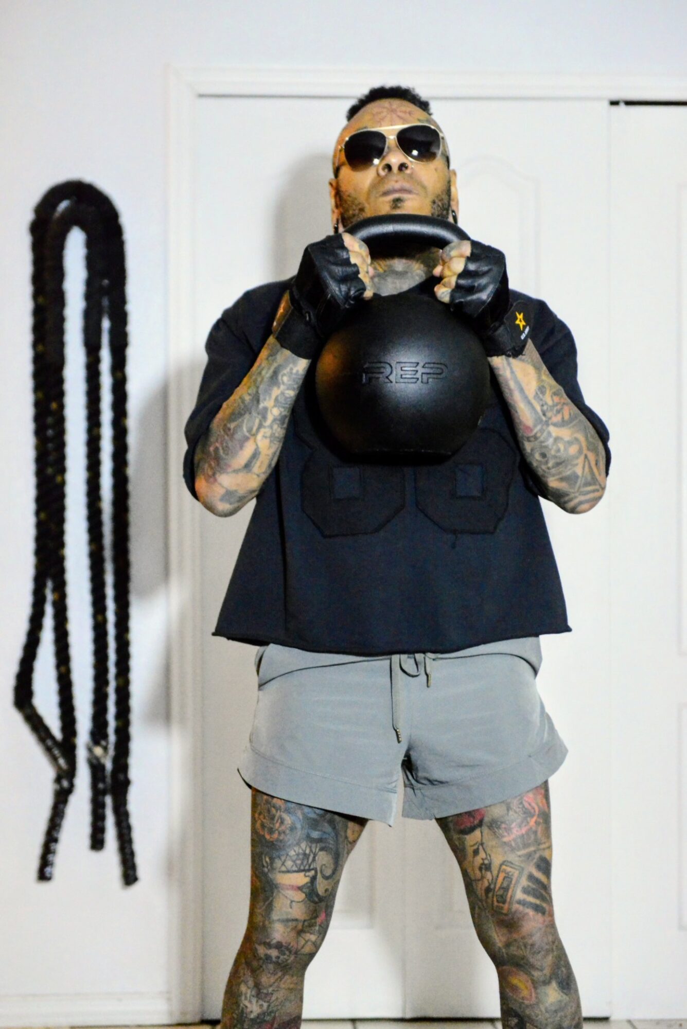 Person with tattoos, sunglasses, black shirt, gray shorts, holding a kettlebell, standing indoors with a rope on the wall behind.