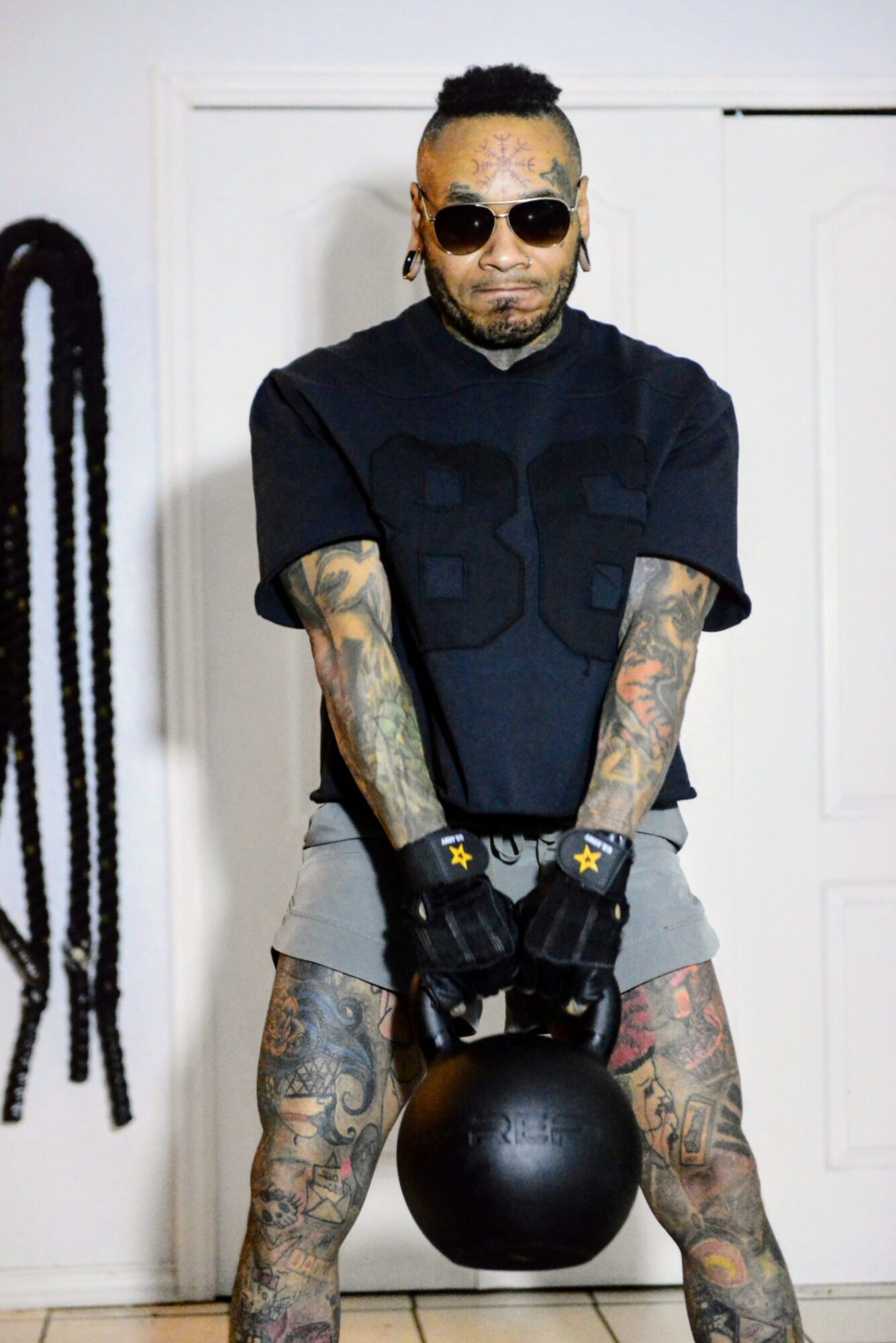 Tattooed man with sunglasses holds a black ball in front of a white door, wearing a black T-shirt and shorts.