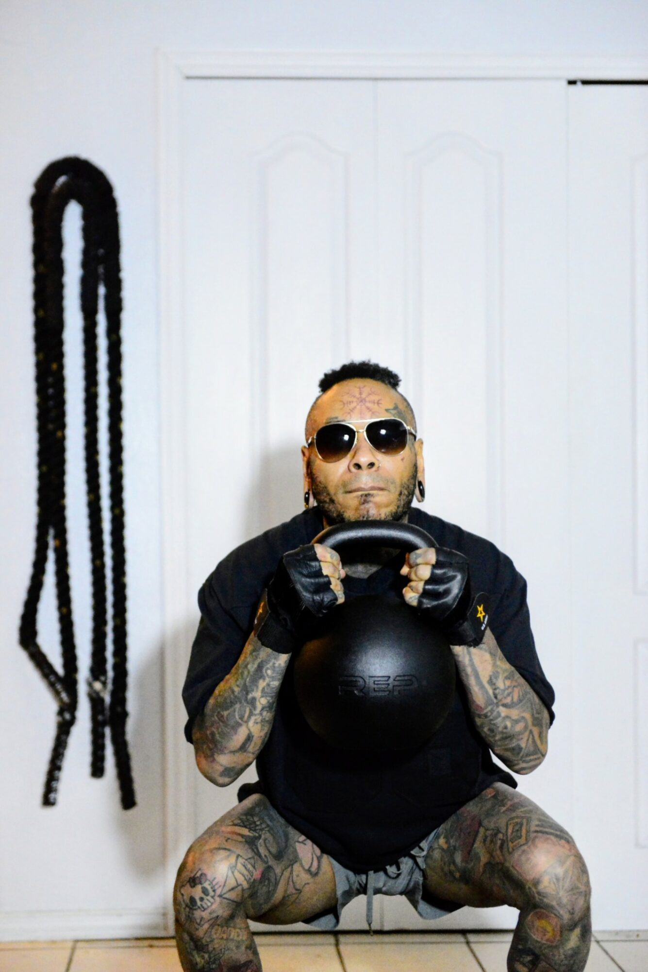 Person with tattoos, sunglasses, and gloves holding a black exercise ball in a room with white walls and a door, with black ropes hanging on the wall.