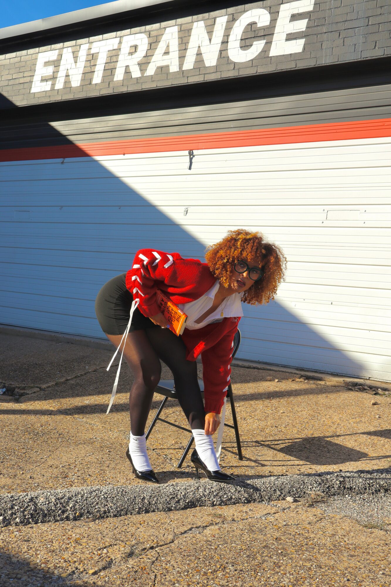 Person with curly hair in red jacket bending down near a white and red garage door outside, with a building and entrance sign above.