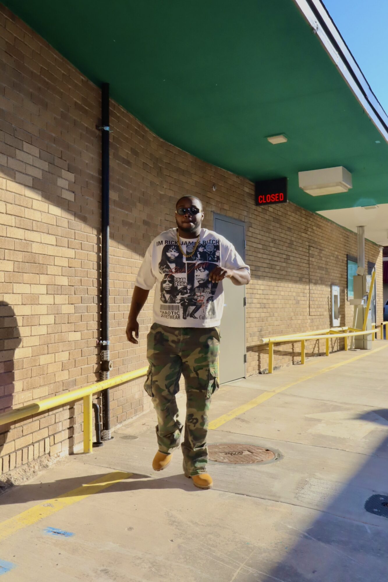 Man walking outside in front of brick wall, wearing sunglasses, a white graphic t-shirt, camouflage pants, and brown shoes.