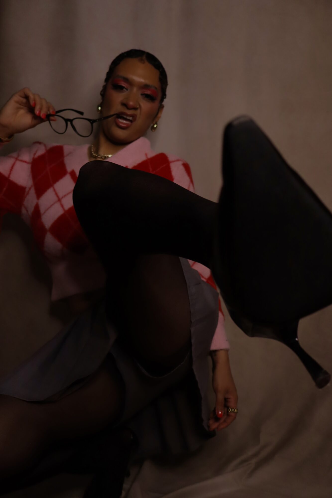 Person with glasses holding them, sitting with legs crossed, wearing a pink and red checkered top, against a plain background.