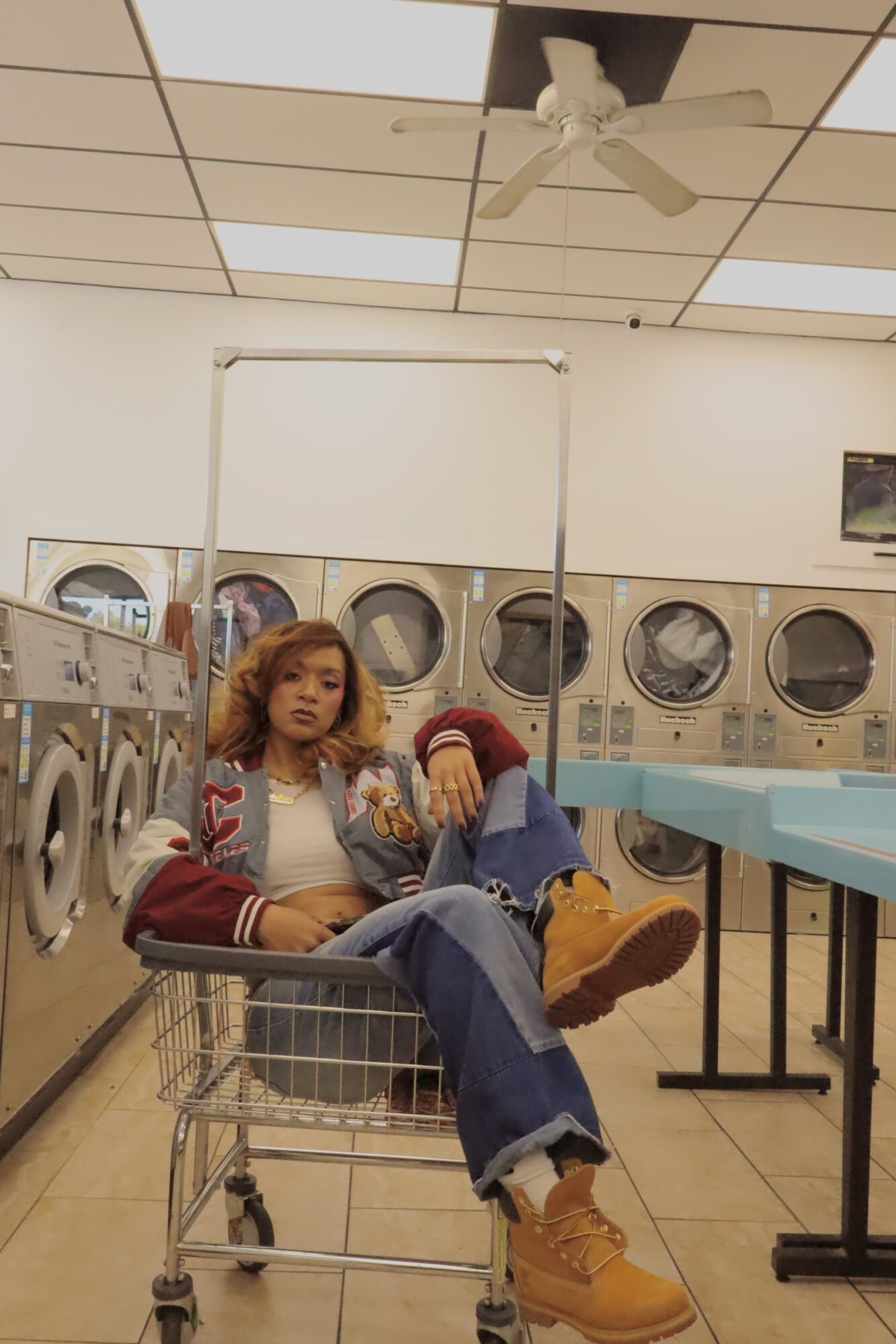 Person with curly hair sitting in a shopping cart inside a laundromat, wearing a jacket, jeans, and boots, with washing machines in background.