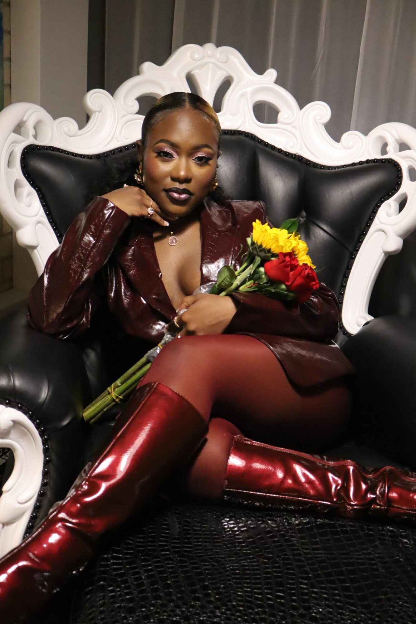 Woman sitting on a black and white ornate chair, holding a bouquet of flowers, wearing a shiny red outfit and dark stockings.