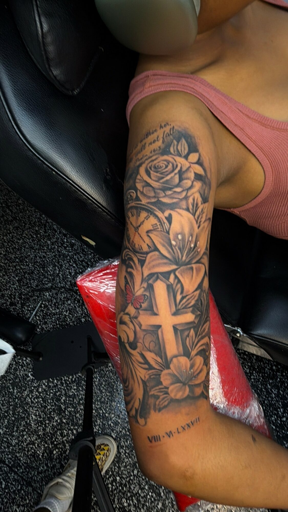 Person with a colorful tattoo sleeve featuring roses, a cross, and text on their arm, wearing a pink sleeveless top.