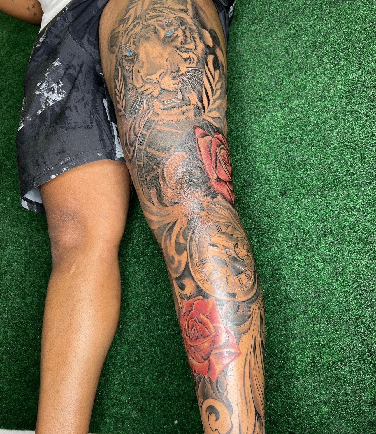 Person with a detailed tattoo sleeve on their leg, featuring a lion, roses, and intricate patterns, lying on green surface.