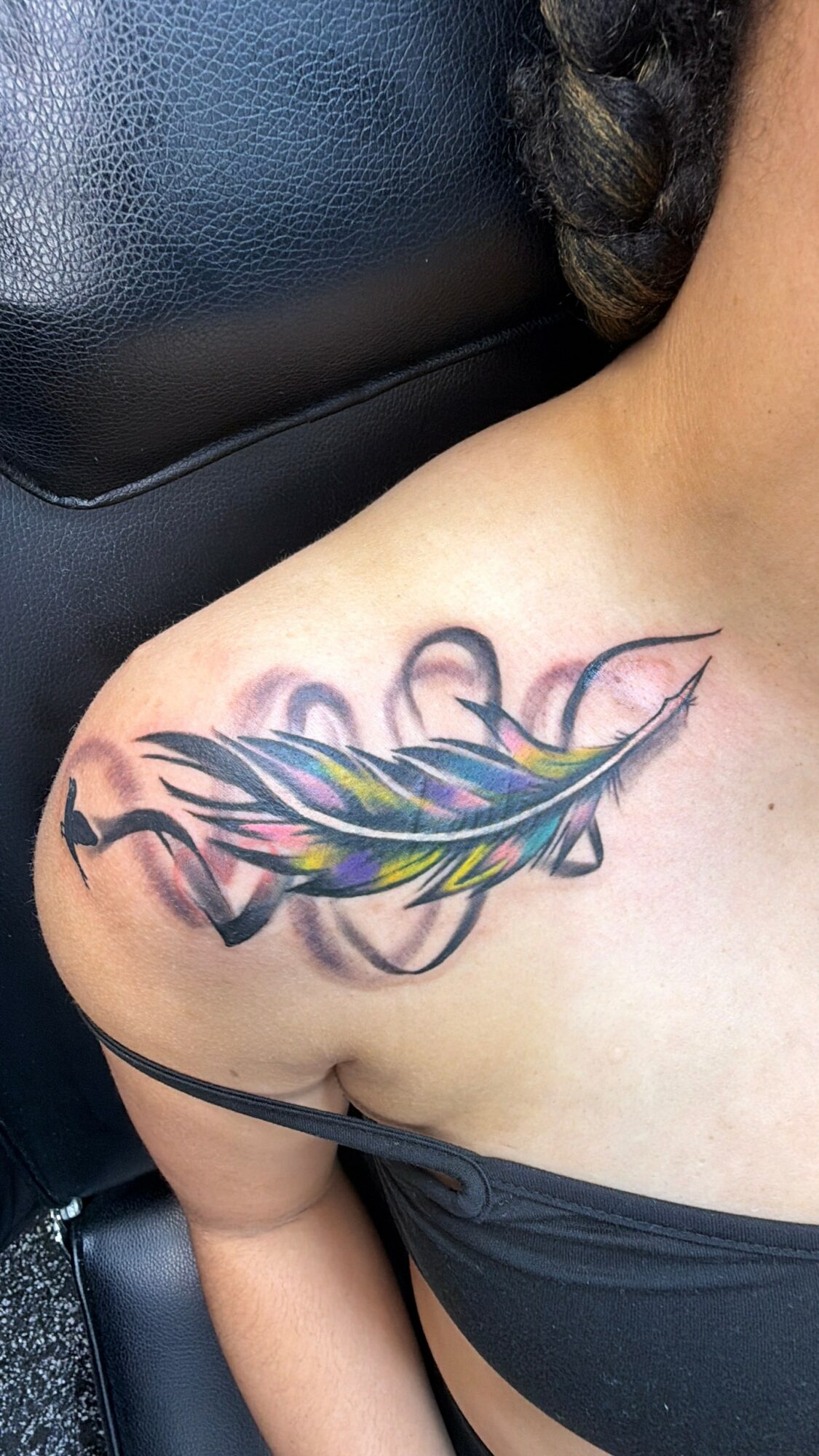 Tattoo of a colorful feather on person's shoulder with black and purple shading, partially visible hair, and black clothing.