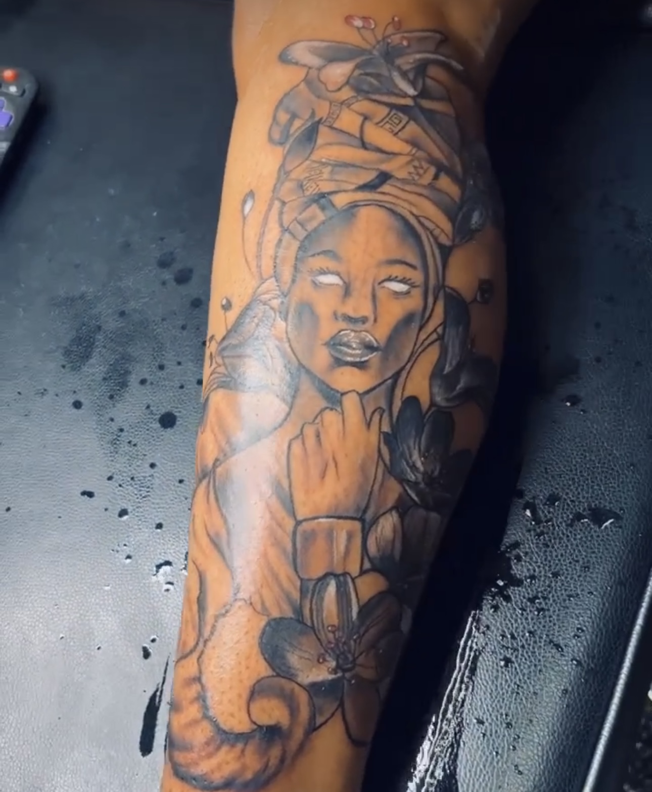 Tattoo of a woman with a headwrap, closed eyes, and flowers on arm, in black and gray ink.