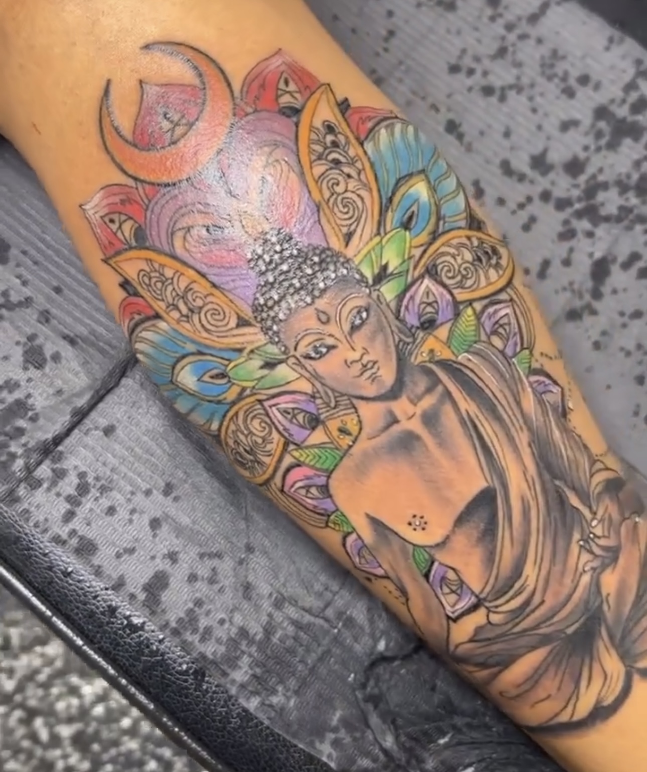 Tattoo of a Buddha figure with colorful, intricate patterns and a moon in the background on a person's arm.