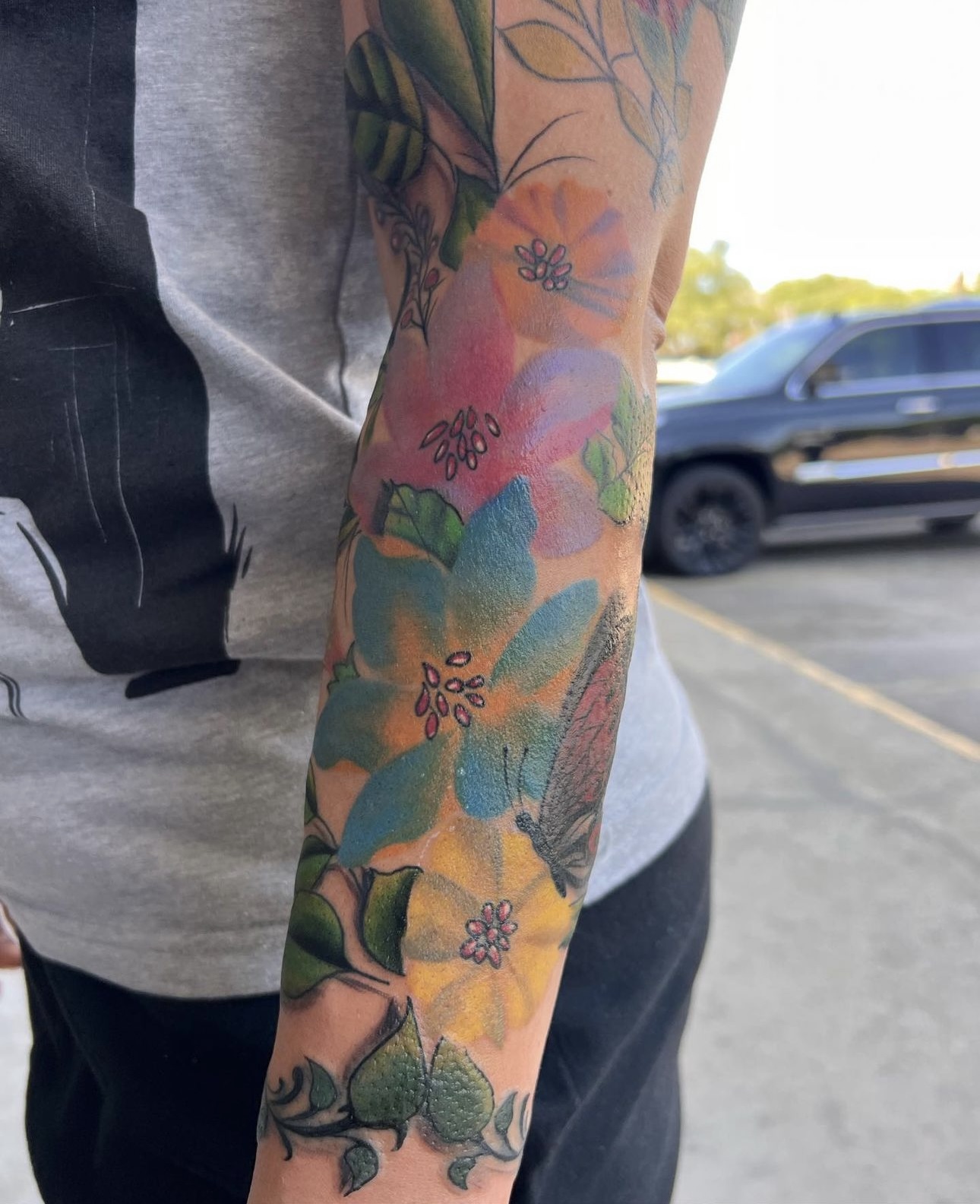 Person's arm with colorful floral tattoo, background shows parked cars and a clear sky.