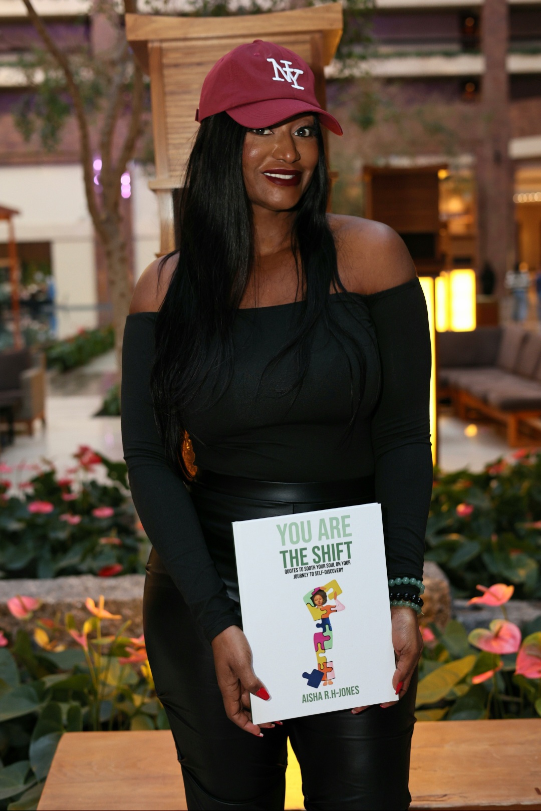 Woman in black dress holding a book outdoors, wearing a red cap, smiling, surrounded by flowers and outdoor furniture.