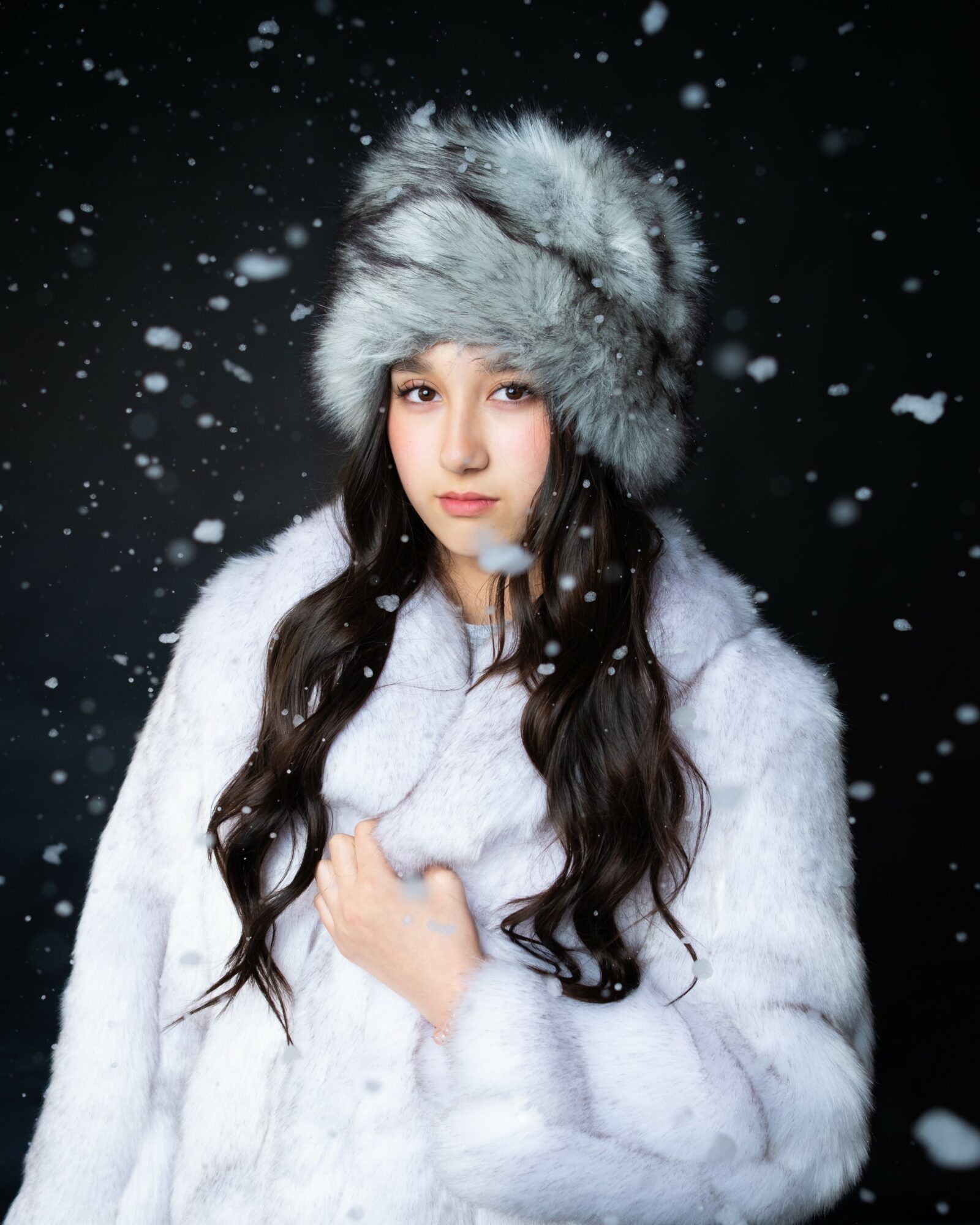 Young woman in a fur hat and coat, with falling snow around her, looking at the camera.