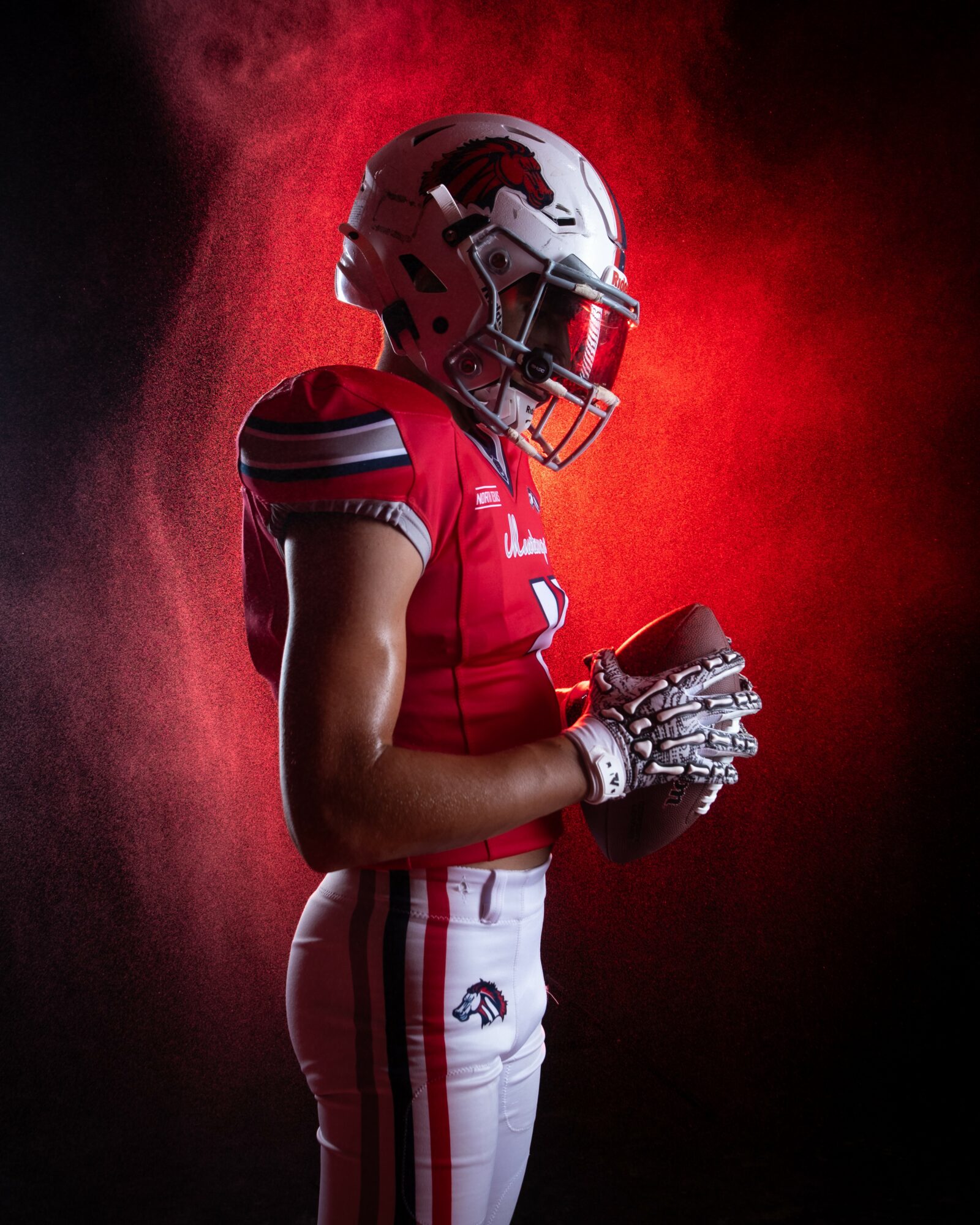 Football player in red uniform and helmet holding a football, standing against a red smoky background.