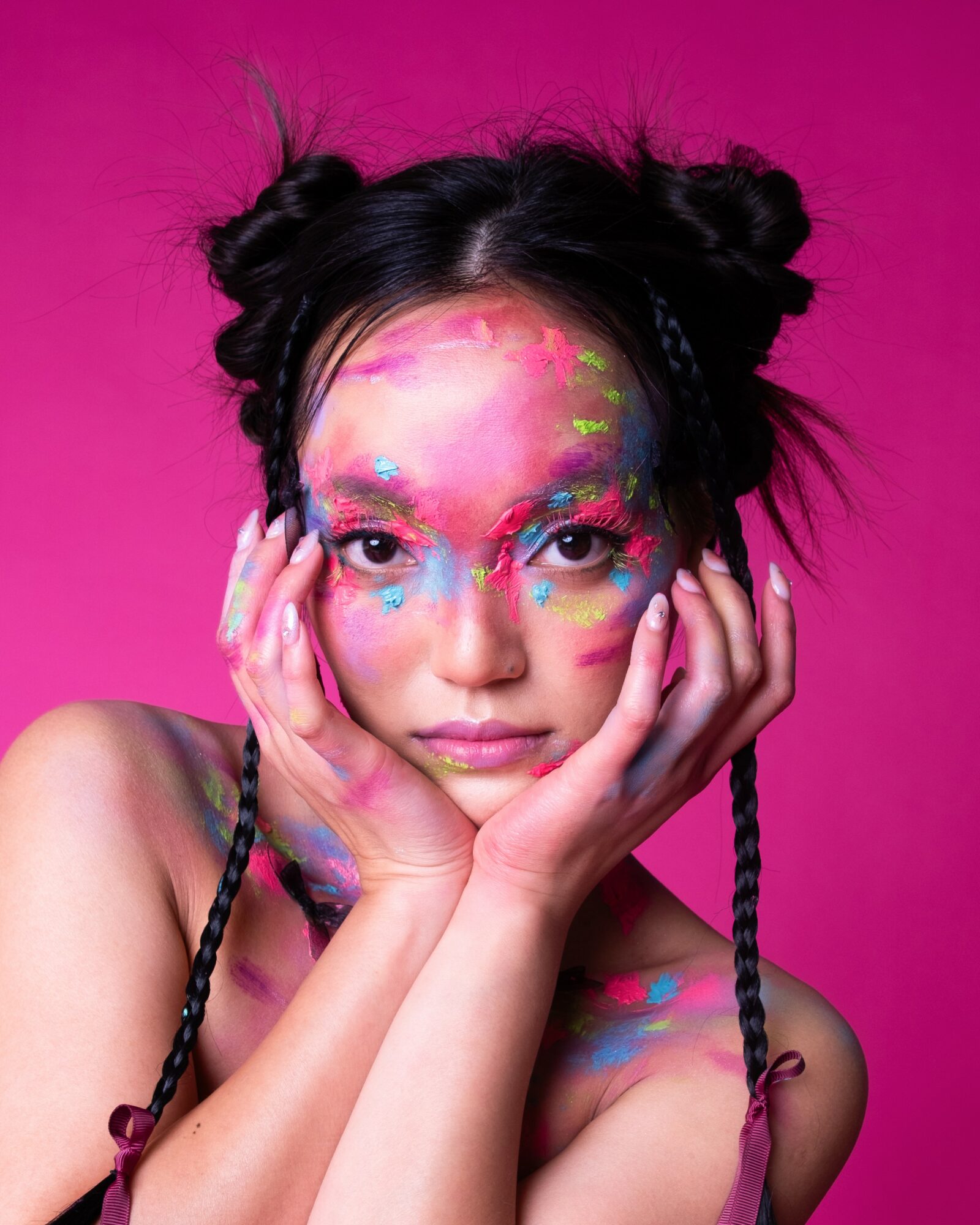 Young woman with colorful face paint and makeup, holding her face with both hands, against a pink background.