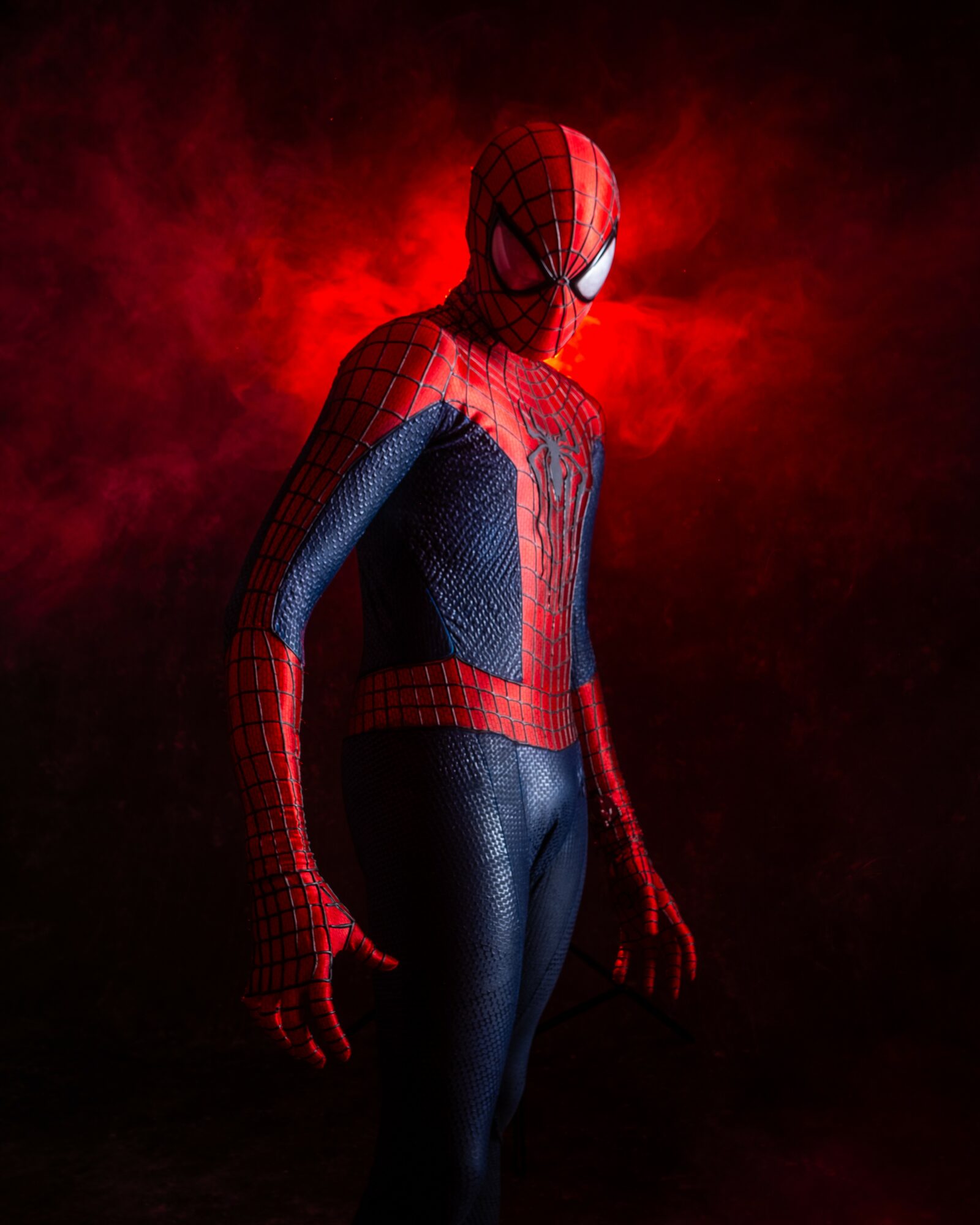 Person in Spider-Man costume standing against a red smoky background, facing slightly to the side with head turned towards the camera.