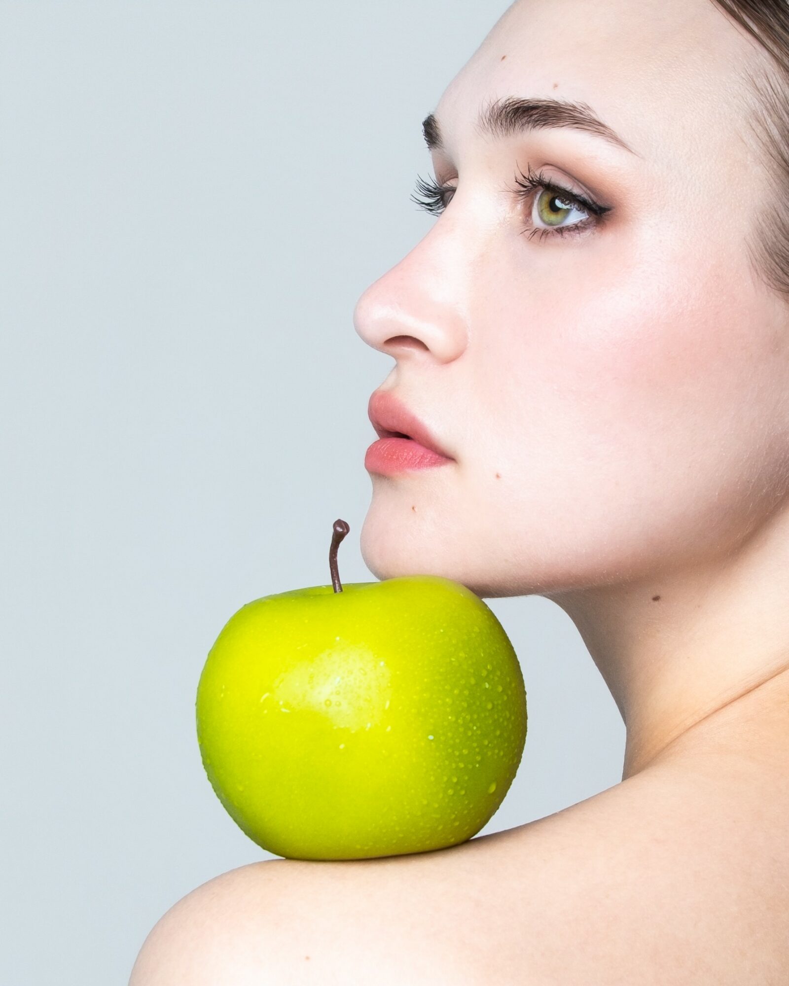 Profile of a woman with green eyes holding a green apple against a light background.