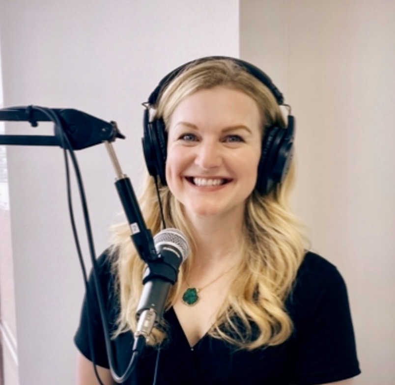 Woman with long blonde hair wearing headphones, smiling, in a recording studio with microphone and pop filter.