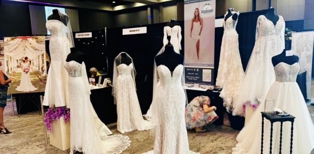 Display of white wedding dresses on mannequins and stands at a fashion exhibition, with posters and other dresses in the background.
