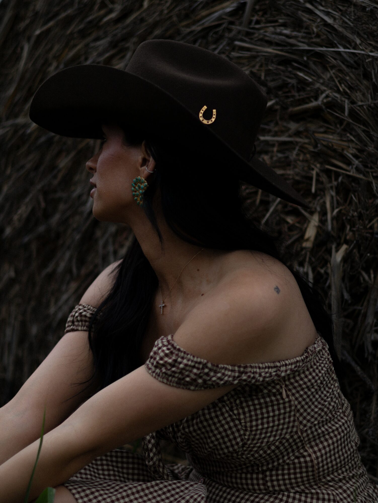 Woman wearing a large hat and off-shoulder dress, sitting outdoors with dark background.