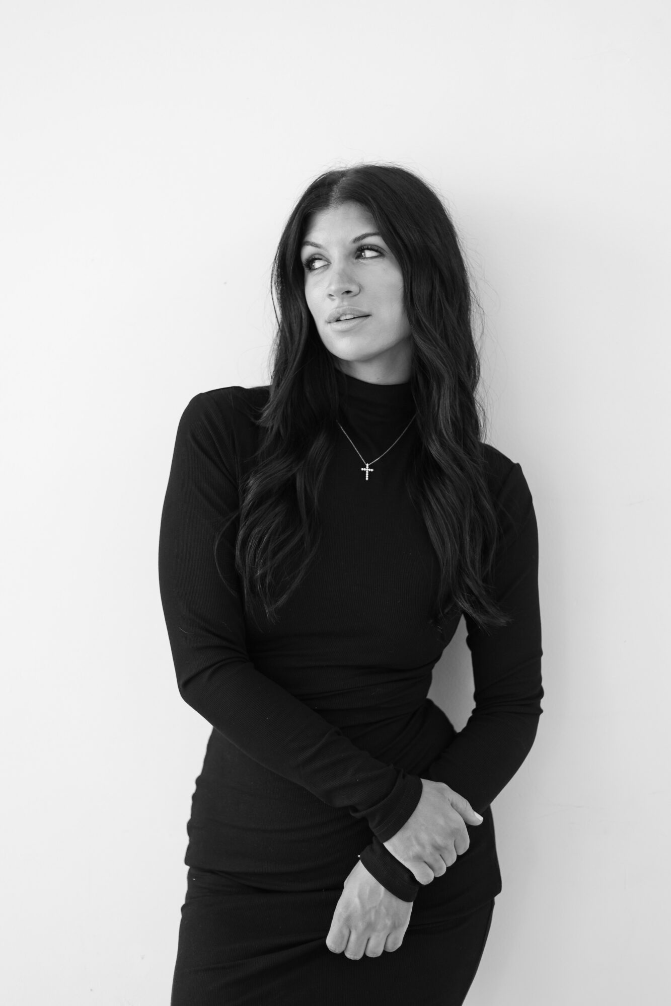 Woman with long dark hair wearing a black long-sleeve top and a necklace, looking to the side, standing against a plain background.