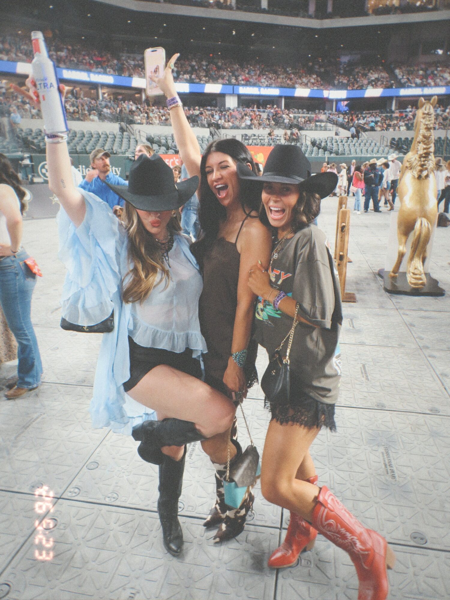 Three women in cowboy hats and boots celebrating at an indoor event, smiling and raising their hands, with a large crowd and a golden horse statue in the background.