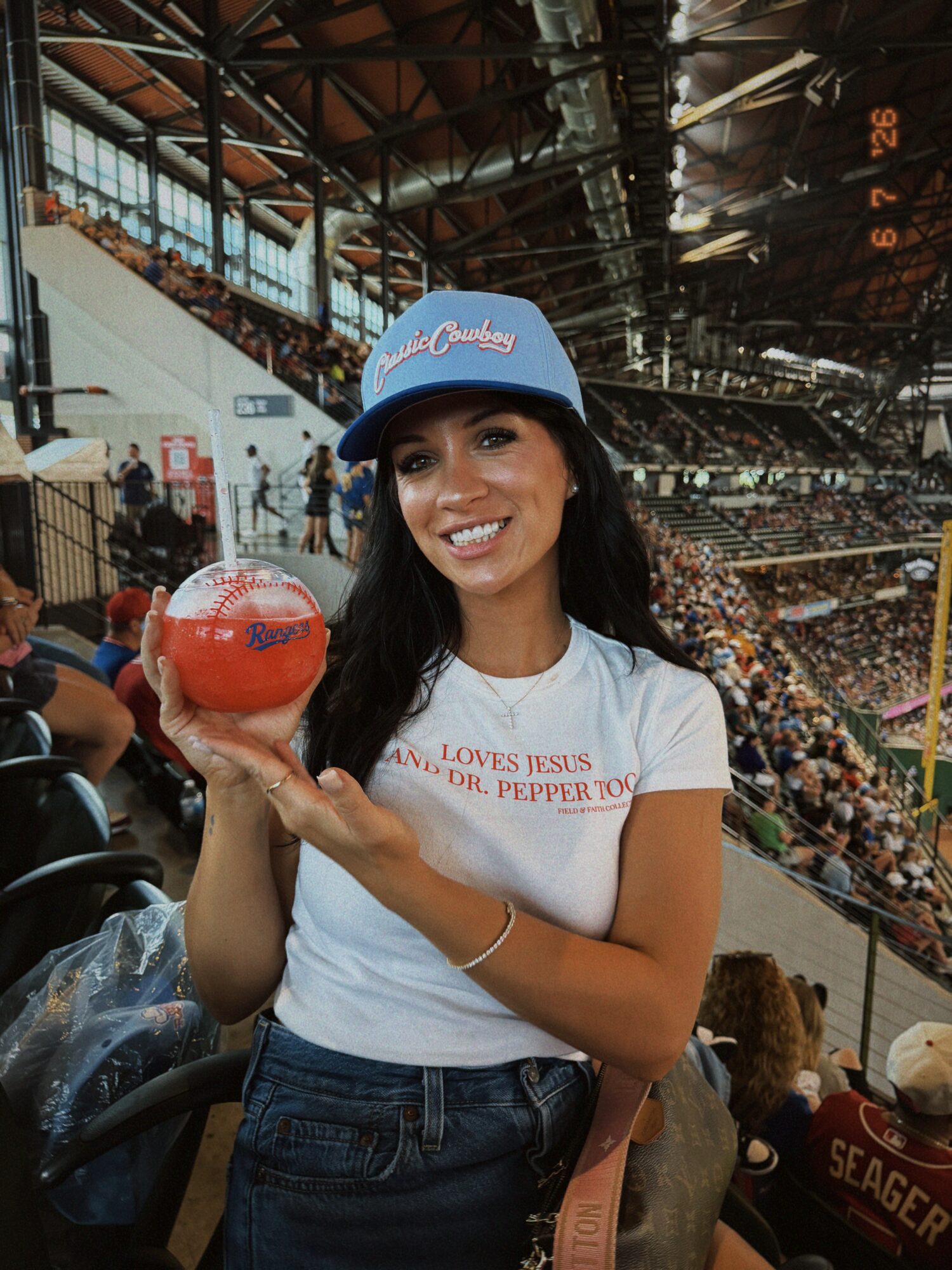 Smiling woman wearing a blue cap and white t-shirt holding a red ball at a stadium.