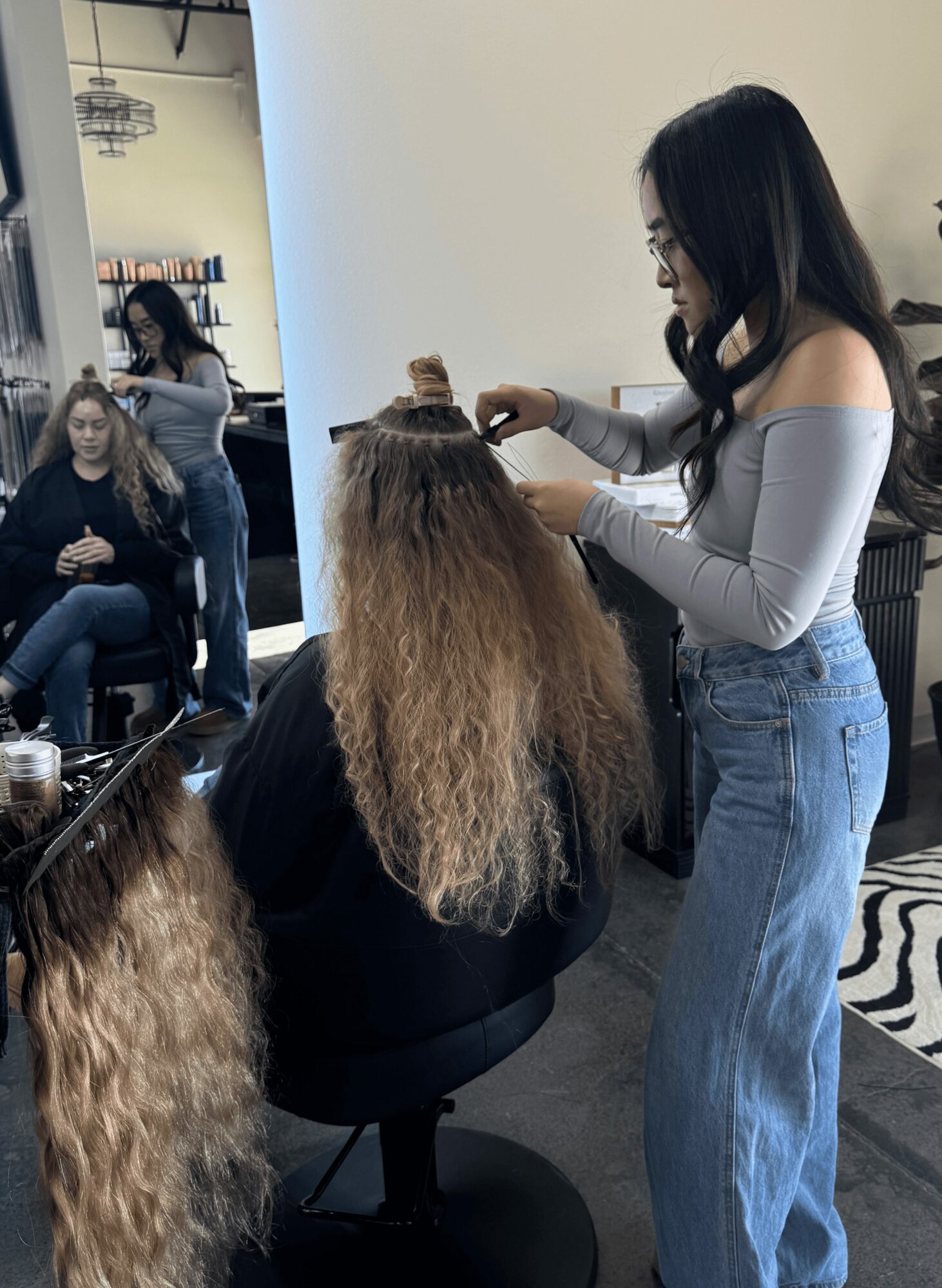 Woman styling a person's long, curly hair in a salon with two seated clients in the background.