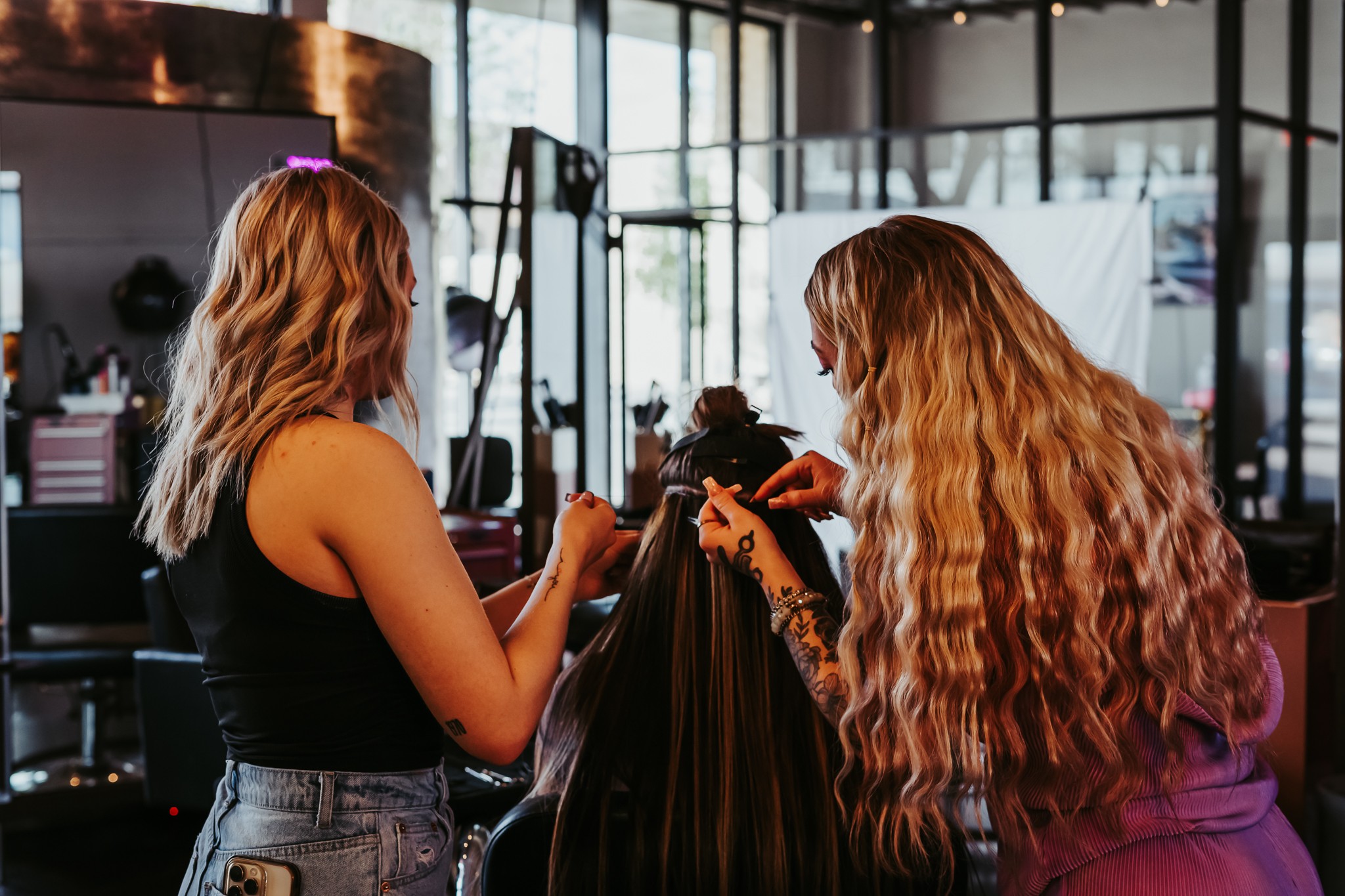 Two women with long, wavy hair in a salon, one styling the other's hair, with salon equipment in the background.