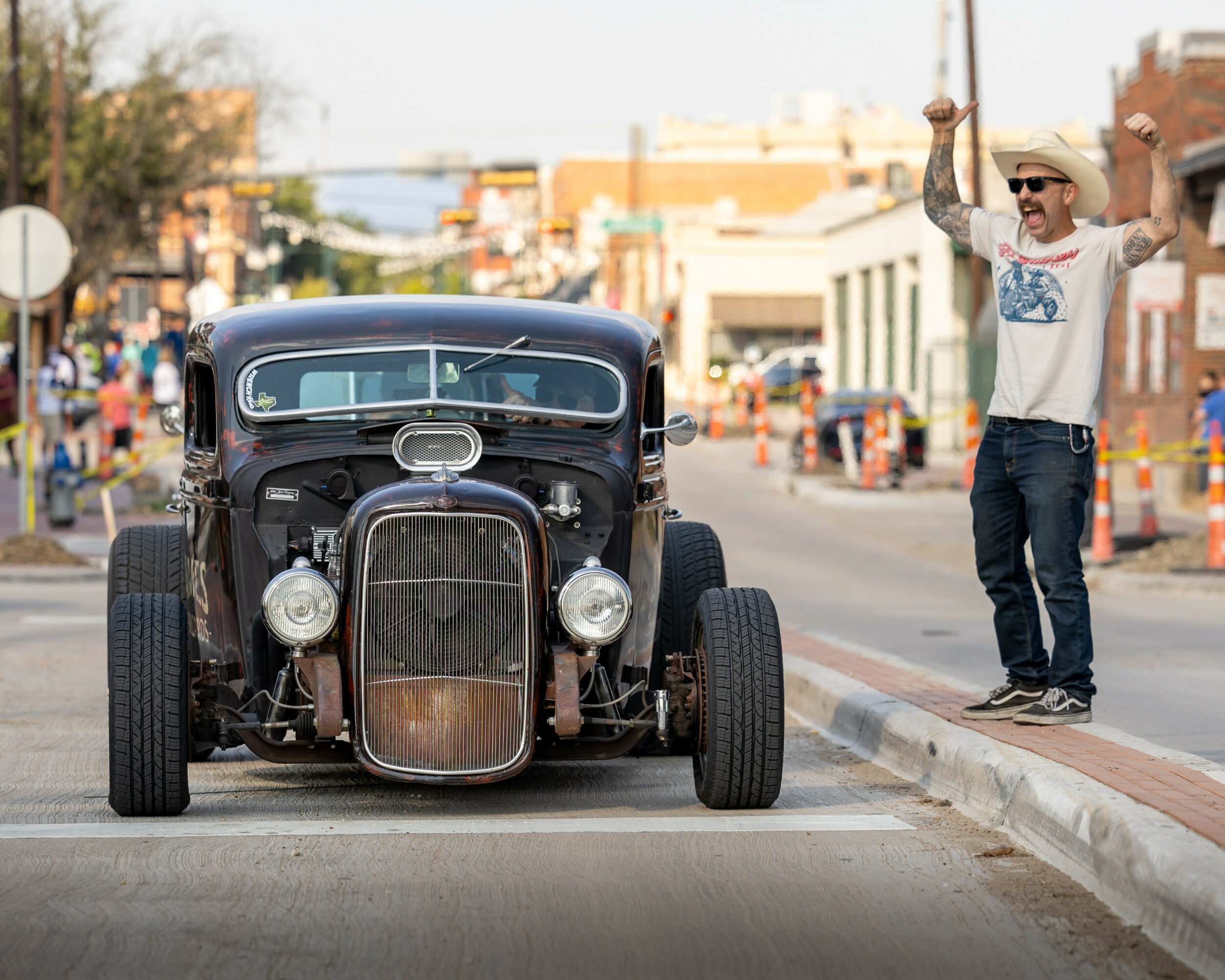 Person standing on sidewalk holding a camera, vintage car parked on street, city buildings in background.