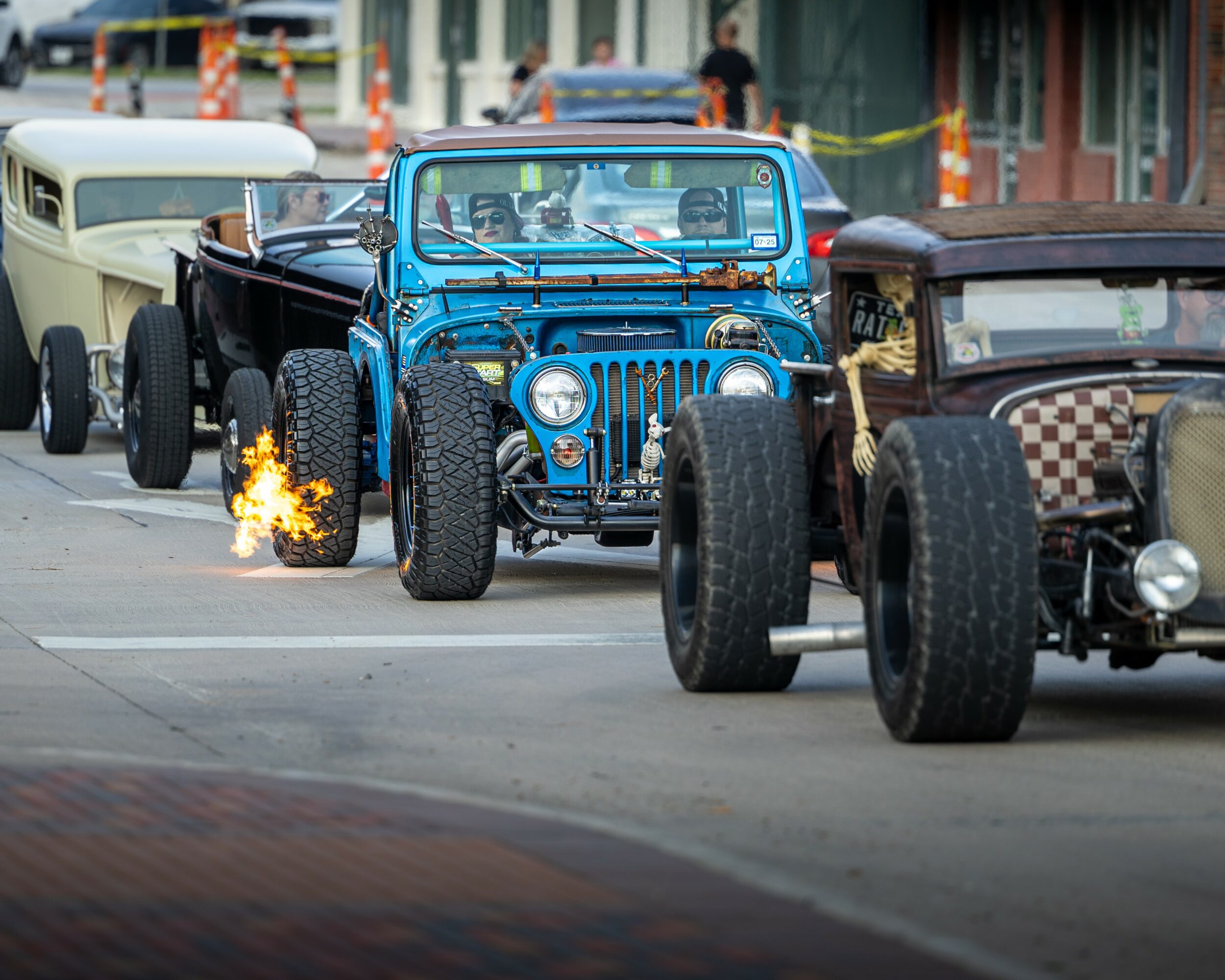 Three vintage cars parked on a street, with one emitting sparks from its wheel, and buildings in the background.