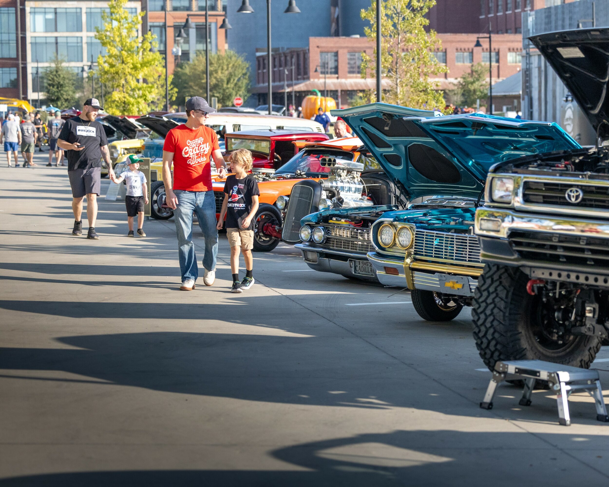 People walking past classic cars on display at an outdoor car show, with buildings and trees in the background.