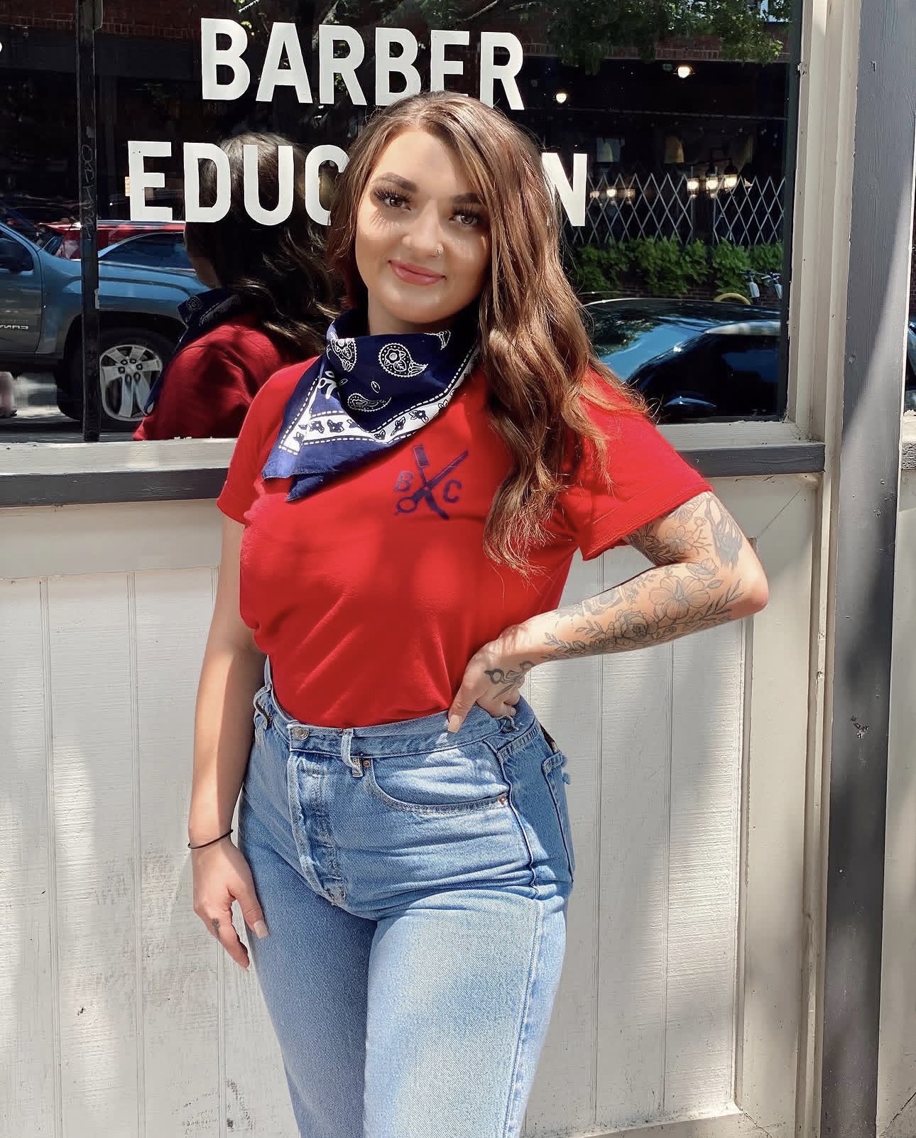 Woman with long wavy hair wearing a red shirt and blue jeans, standing outside a building with a window.