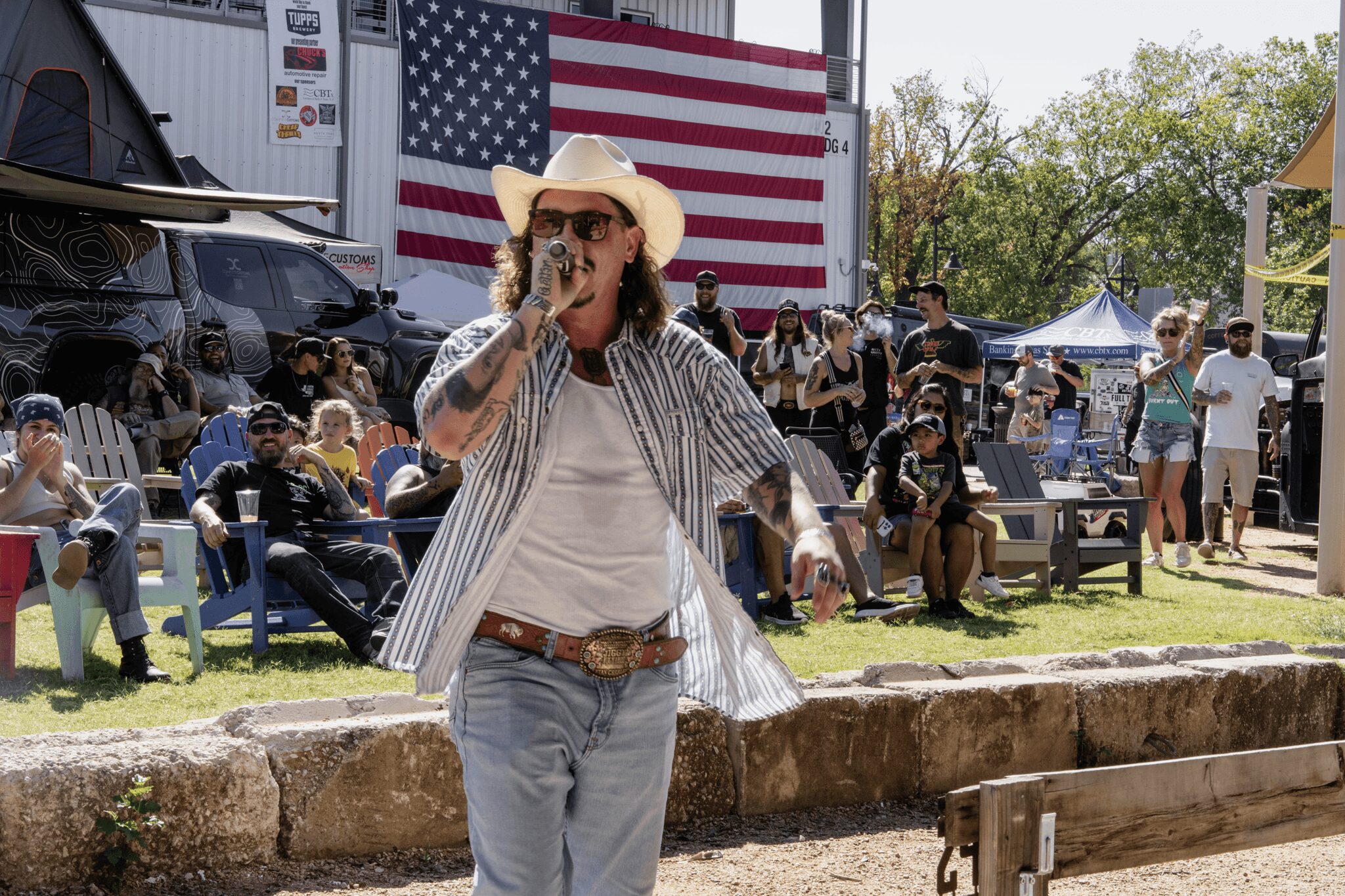 Person wearing a hat and sunglasses, holding a microphone, standing outdoors with a crowd and American flag in background.