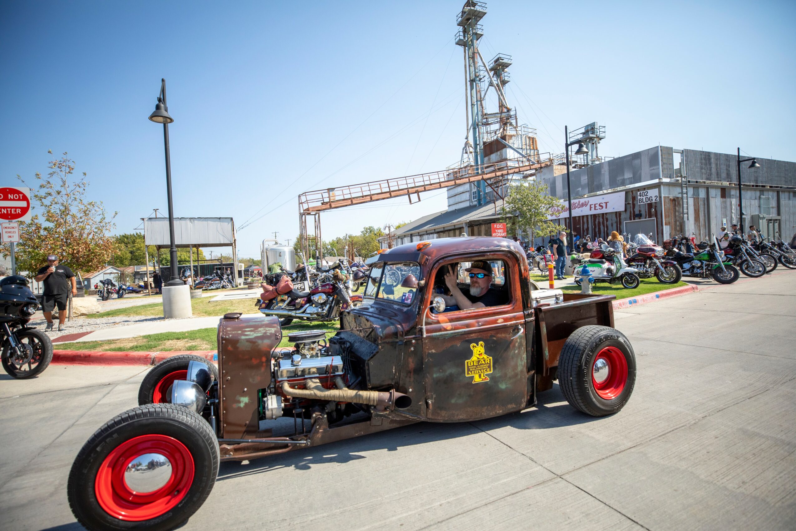 Vintage car with exposed engine and red wheels driving on street, with motorcycles and industrial buildings in background.