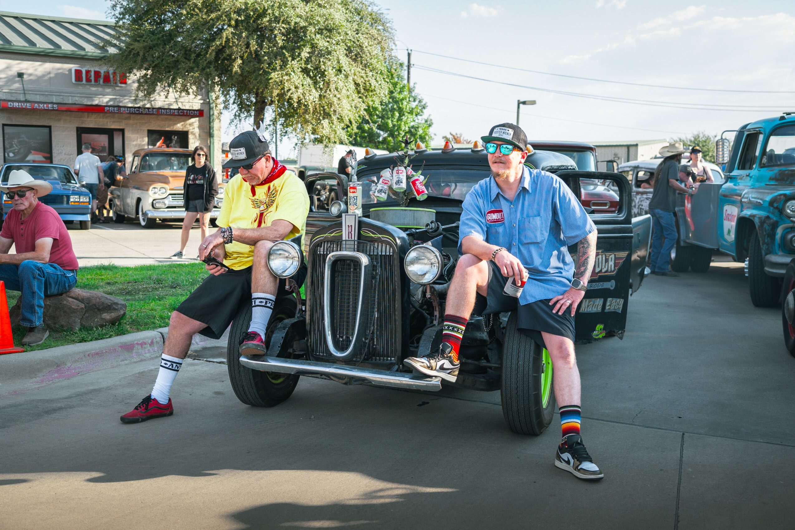 Two people sit on a vintage car, one in a yellow shirt and the other in a blue shirt, in a parking lot.