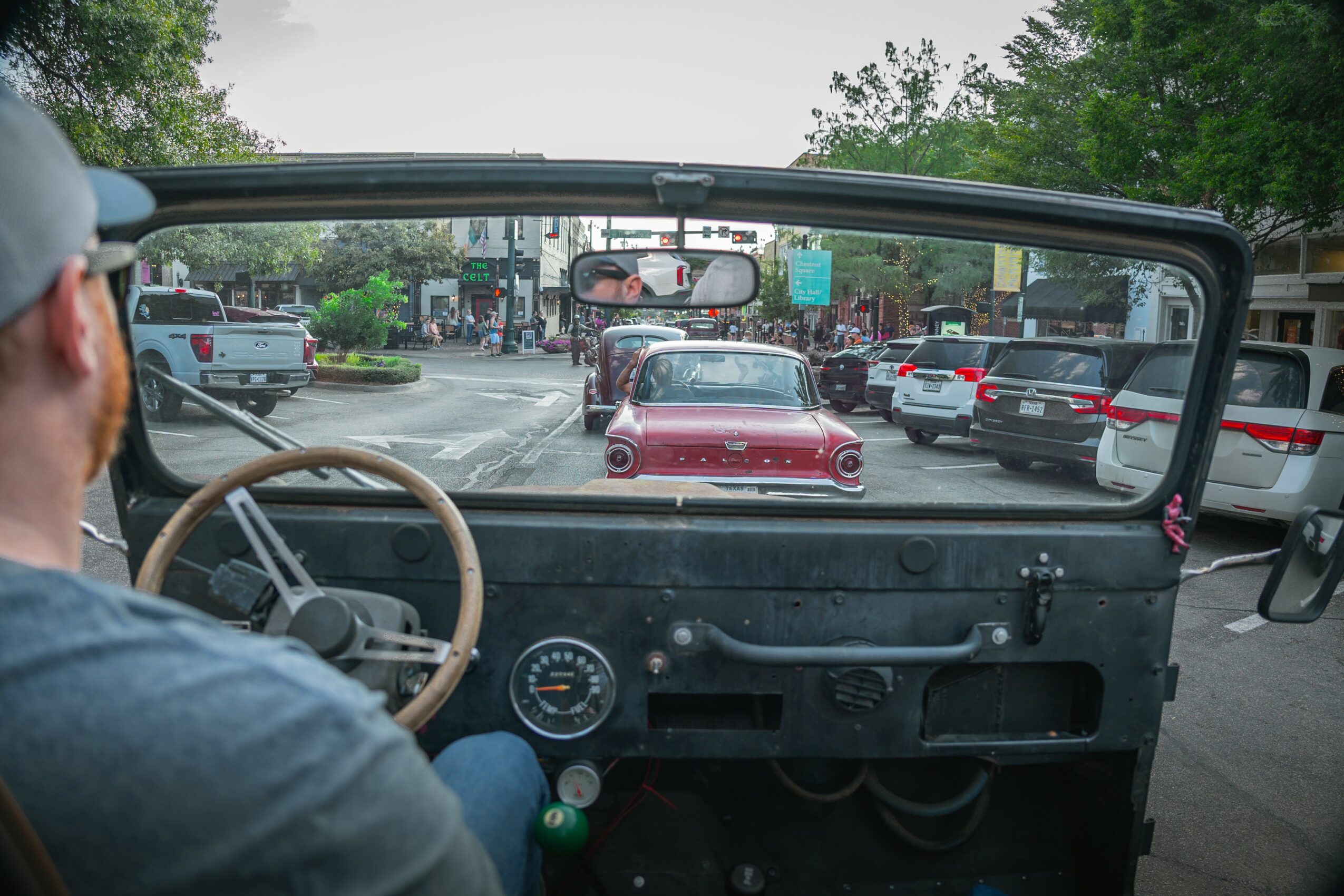 View from inside a vehicle showing a street with cars and trees, driver visible on the left, dashboard in foreground.