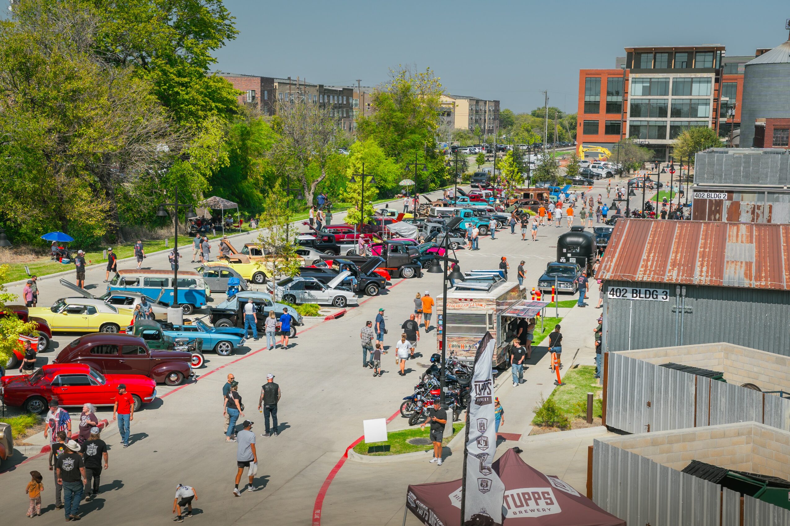 Street scene with parked cars, people walking, trees, and buildings in the background.